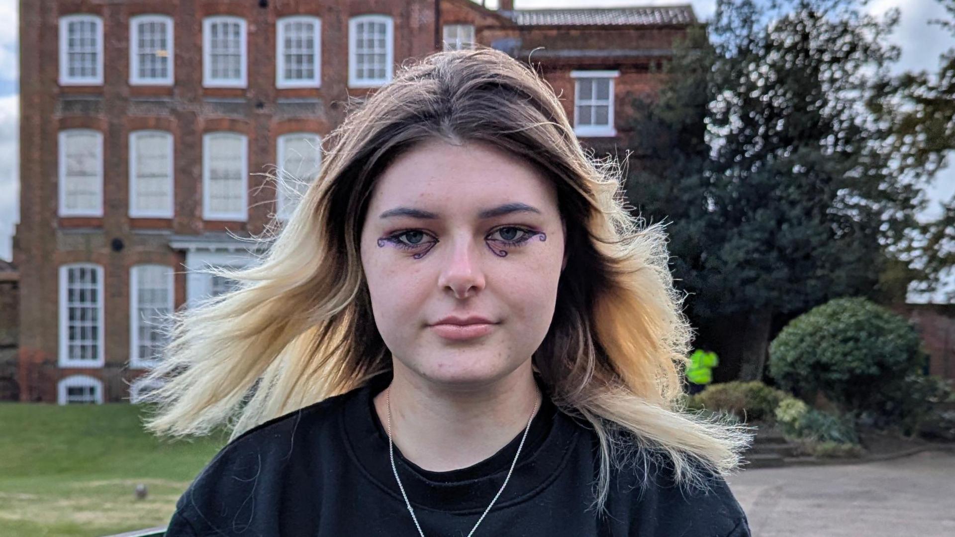 Kaya has blonde hair which is dark at its roots. She is wearing a black jumper and has blue makeup around her eyes. She is sitting in front of Hollytrees Museum in Colchester, which is a 18th Century brick townhouse.