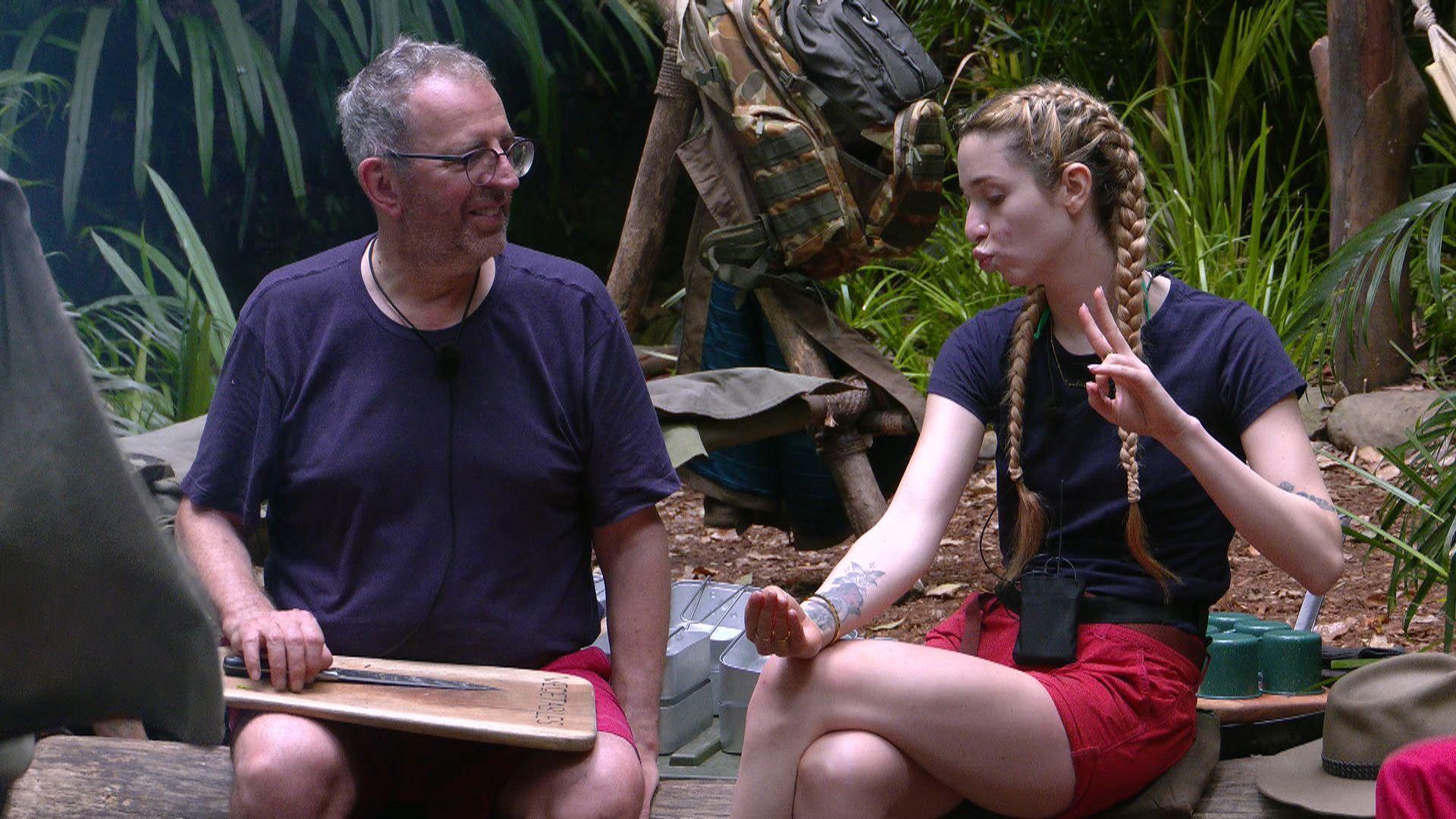 Rev. Richard Coles wearing a blue t-shirt, holding a chopping board and knife watches GK Barry pouting and holding her fingers in a peace sign