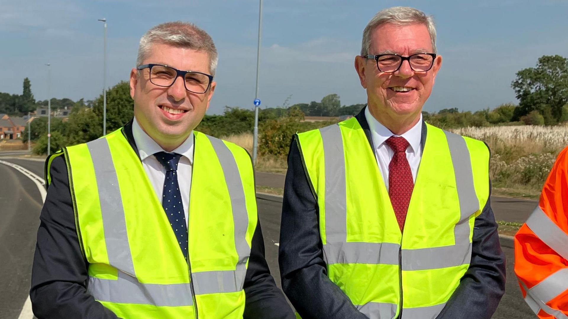 A man in a navy tie and green high visibility jacket stands next to a man in a red tie and green high visibility jacket standing in a road.