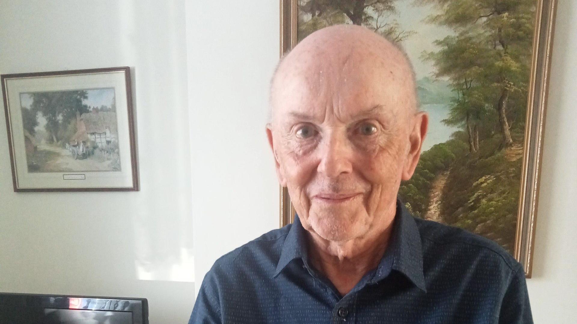 Barry Watson, an elderly man wearing a dark blue shirt looks at the camera and smiles. He appears to be in his house standing in front of a painting. 