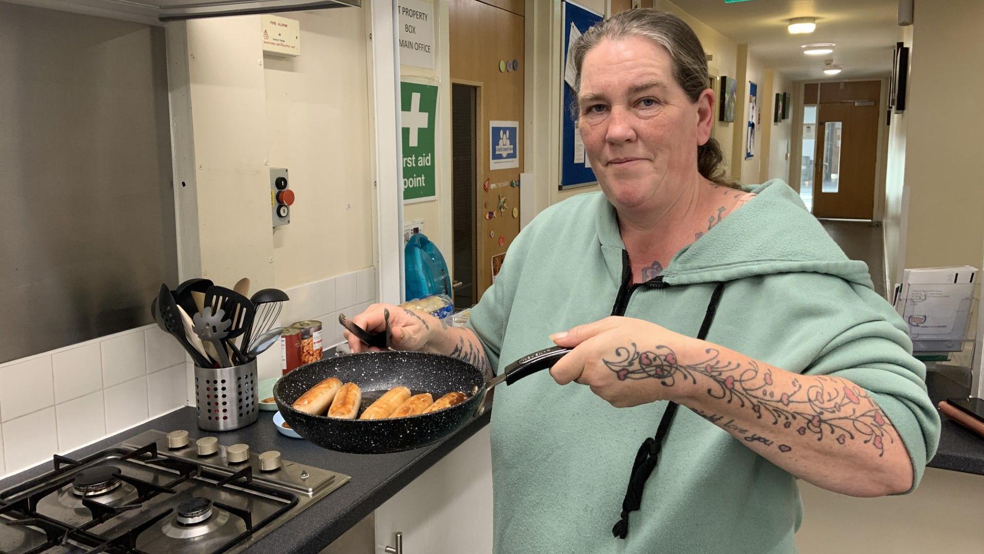 A woman is stood in a kitchen at a community centre. Her hair is tied back and she is wearing a green hoodie. She is holding up a frying pan full of sausages