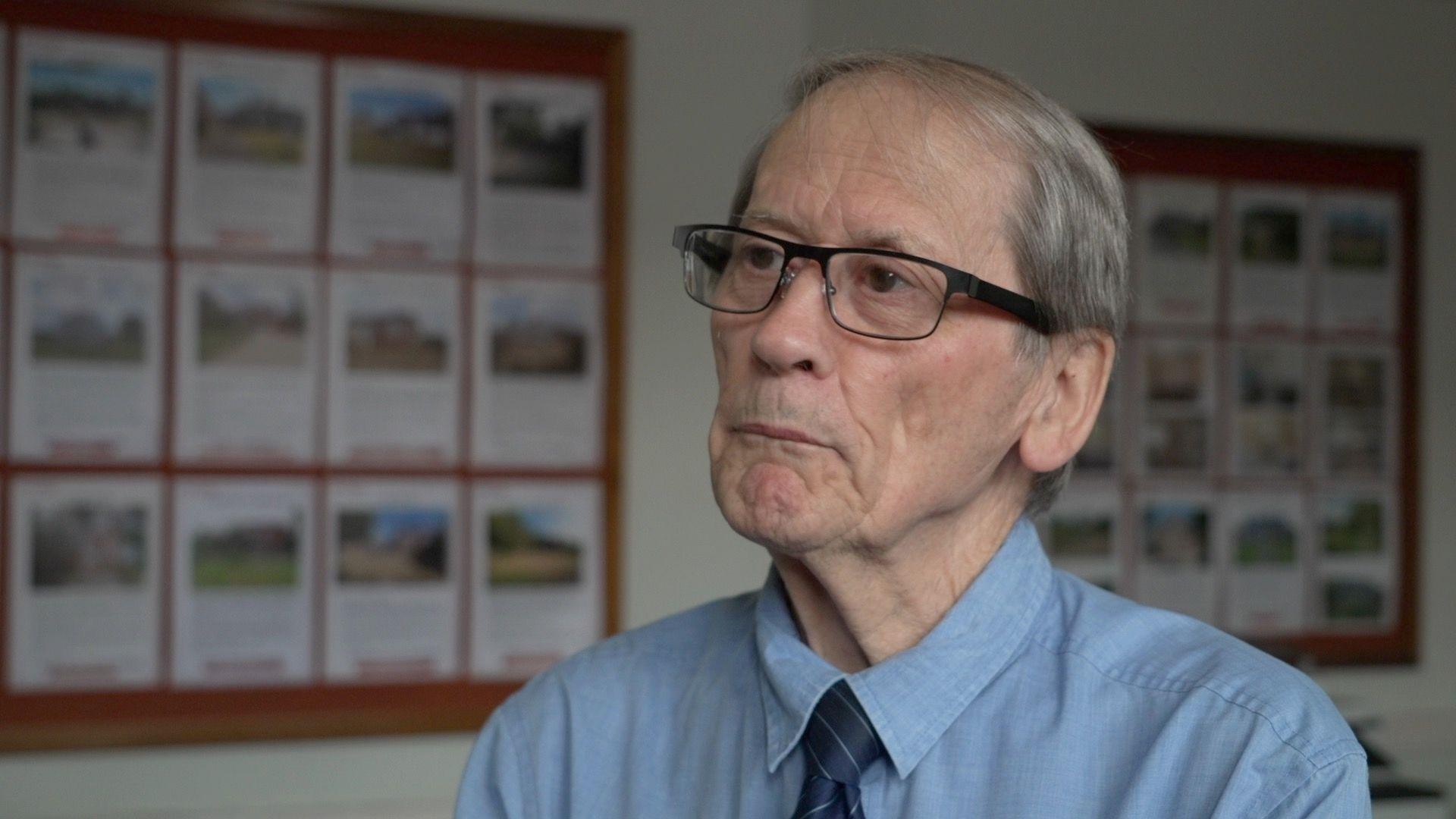 A man with grey hair and black-framed glasses stands in front of a board of house adverts in an estate agents.