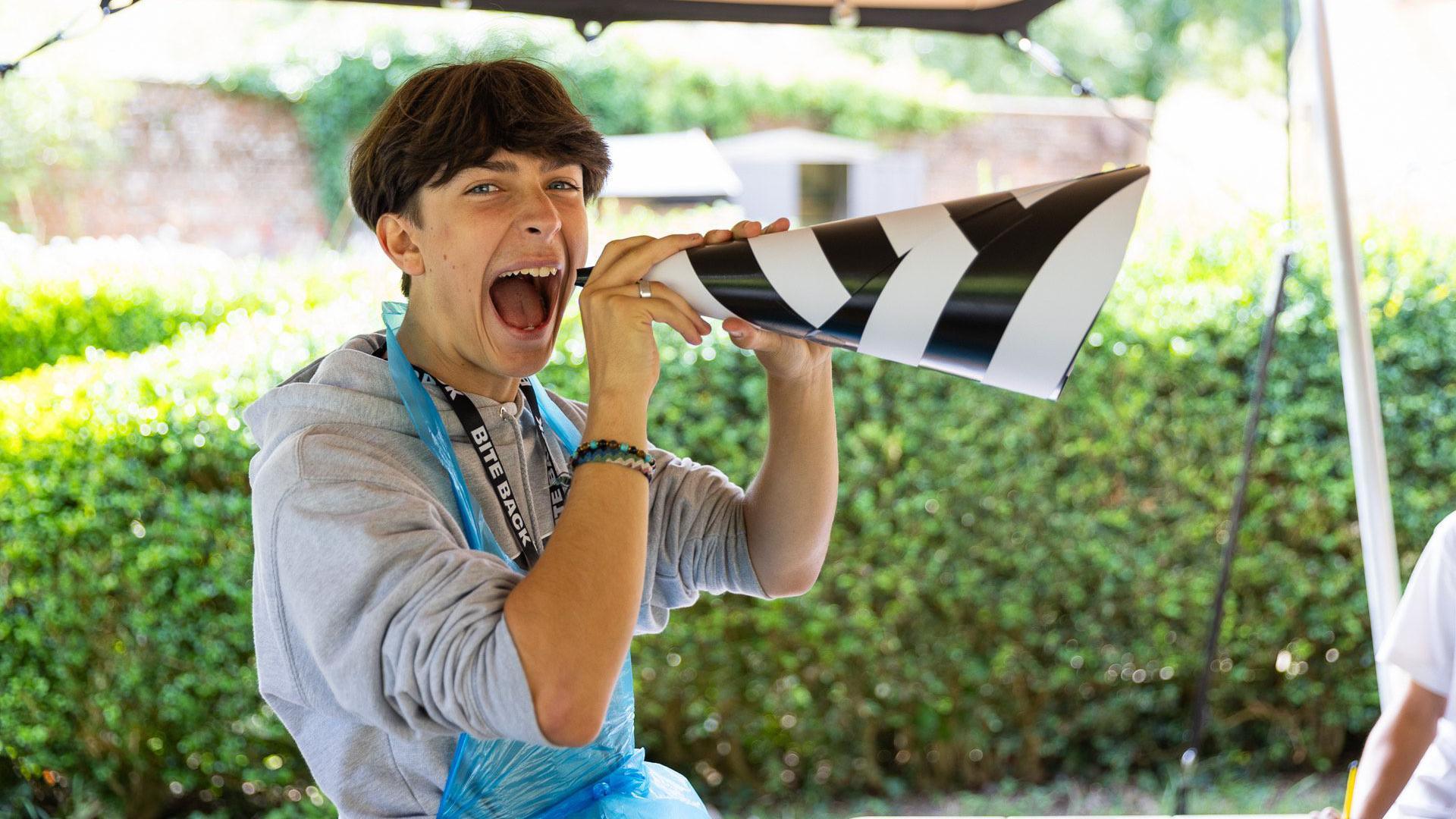 A man holding a black and white cone to his face. He has his mouth open. He is stood in front of a green box hedge.