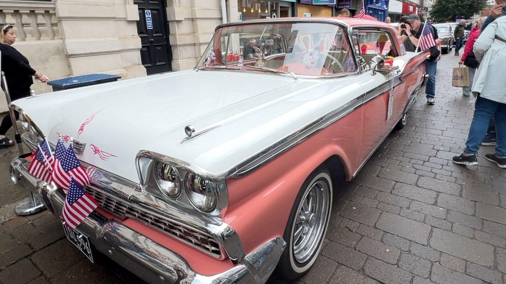 A retro car on the street in Gloucester's city centre. The bodywork is a mixture of salmon pink and white and there are a handful of USA stars and stripes flags attached to the bonnet