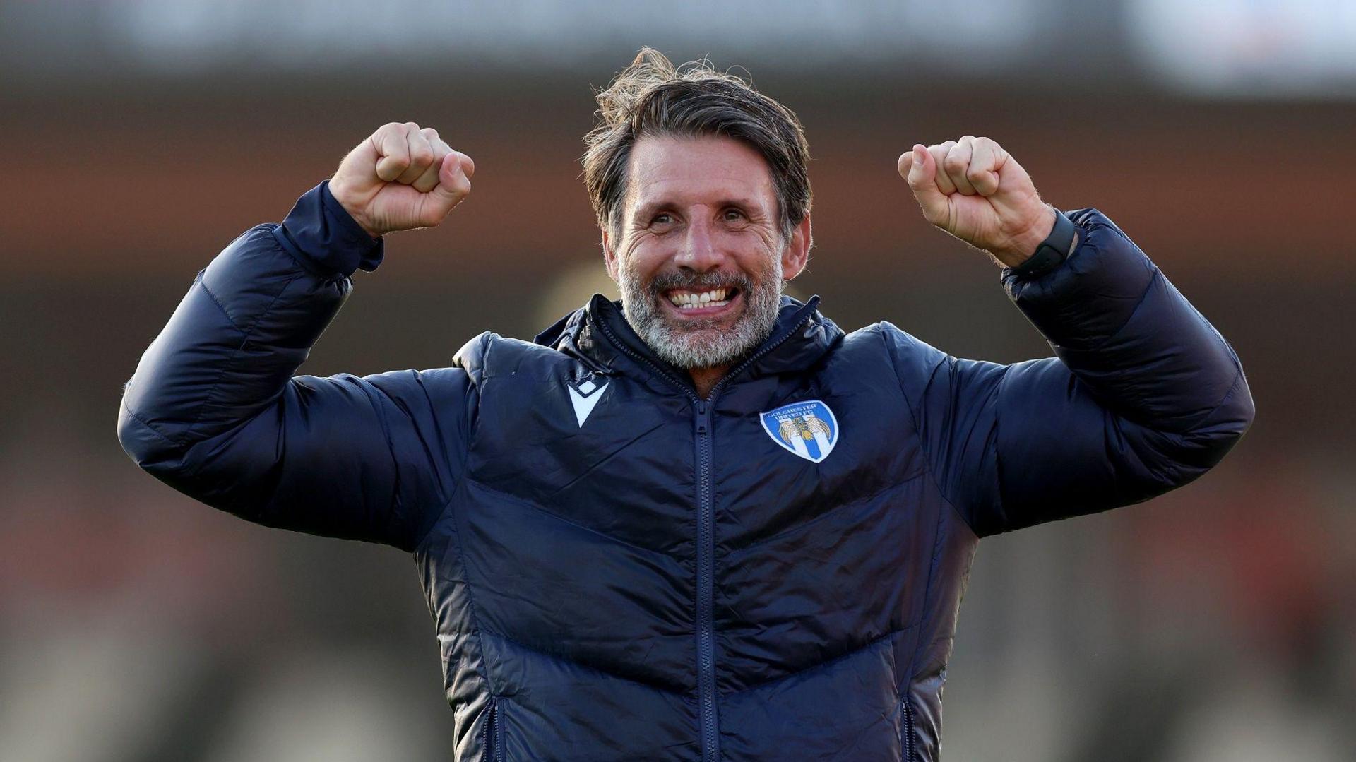 Danny Cowley is wearing a navy tracksuit zip-up top, with the Colchester United crest printed on it. He is raising his arms and clenching his fists in celebration, with a big smile on his face, looking just beyond the camera lens.