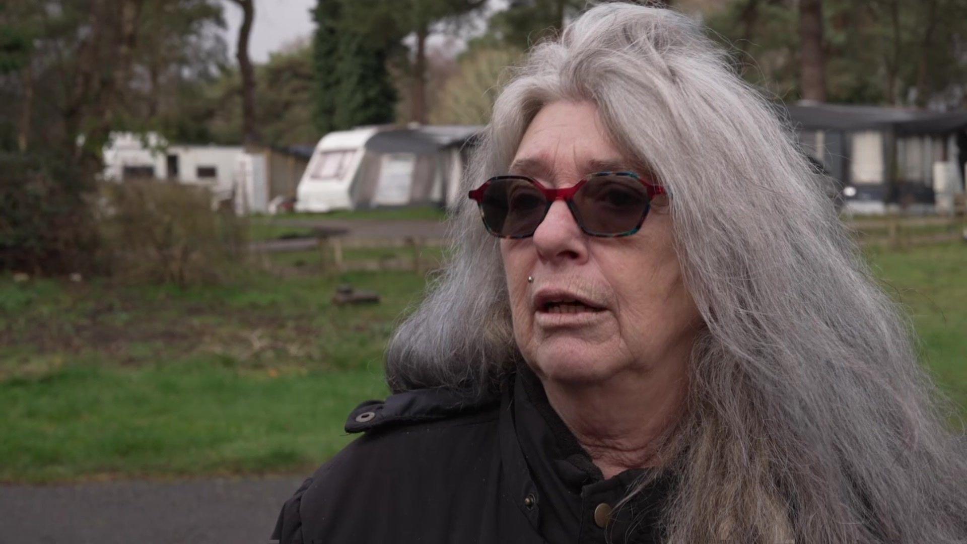 A woman with grey hair and wearing sunglasses looking straight at the camera with a row of caravans behind her.