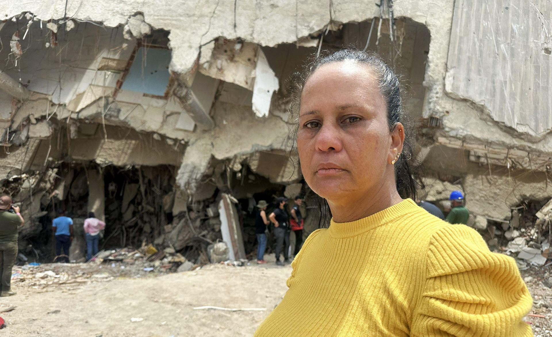 Laura Barrios stands in front of a building as she searches for the remains of her brother-in-law. She is wearing a yellow sweater, her dark hair is pulled back.