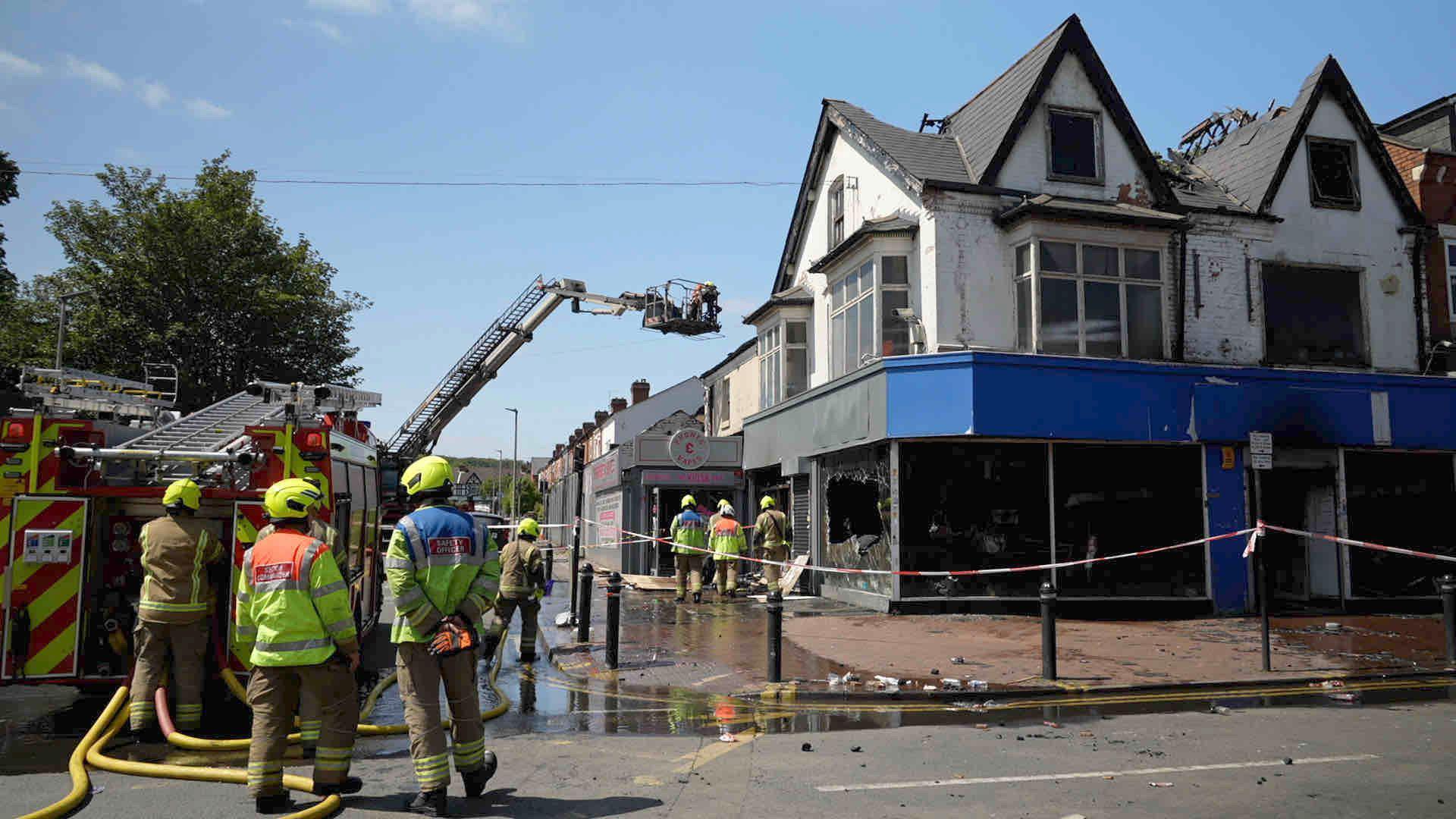 Three fire staff are standing behind a red and yellow fire engine, which is parked in front of a shop. that has blue and black frontage. There is a red and white cordon, and its windows are smashed. There are parts of the roof missing and the first floor is blackened with fire. The fire engine has deployed an aerial ladder which is hovering above the building