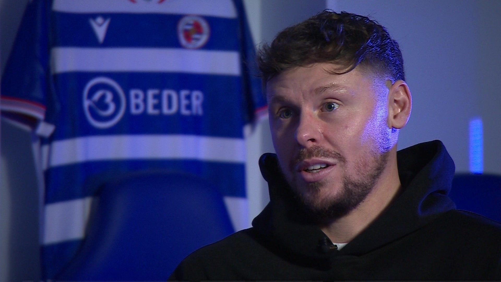 A blonde man wearing a black hoodie who appears to be talking to someone off-camera. There's a blue and white stripy shirt hanging behind him with "Beder" written across the front.