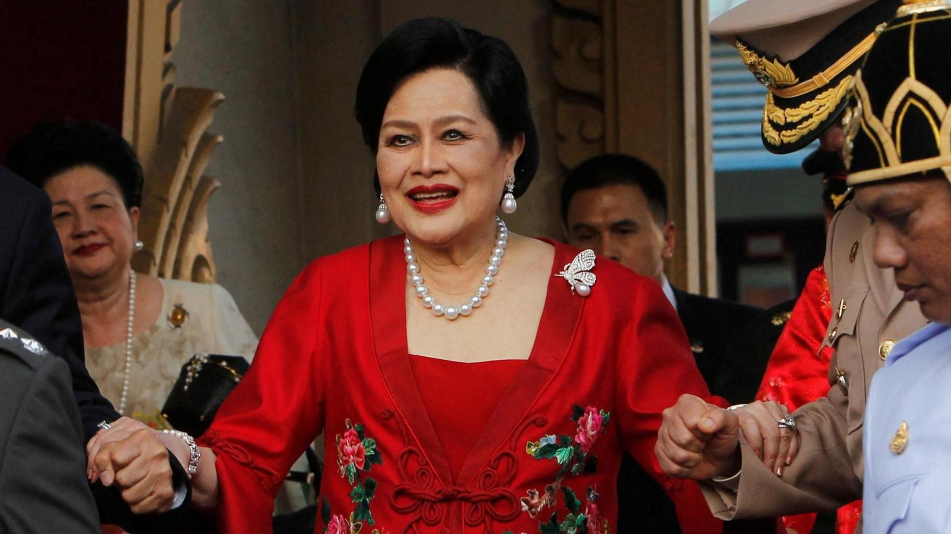 Queen Sirikit, wearing pearl jewellery and a red dress, is helped down a staircase by guards in uniform.