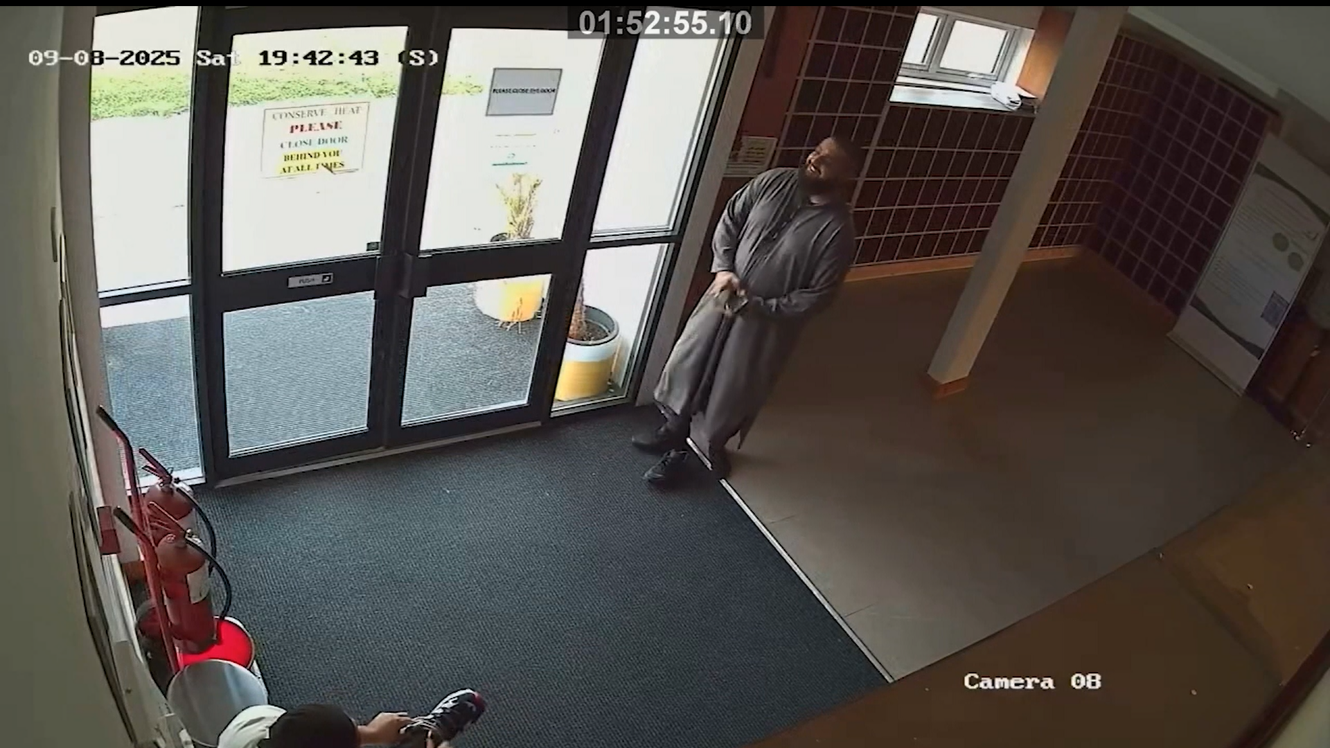 CCTV view of a man in a dark grey outfit smiling by the door of a hallway. Another man sits in the corner putting on a pair of shoes.