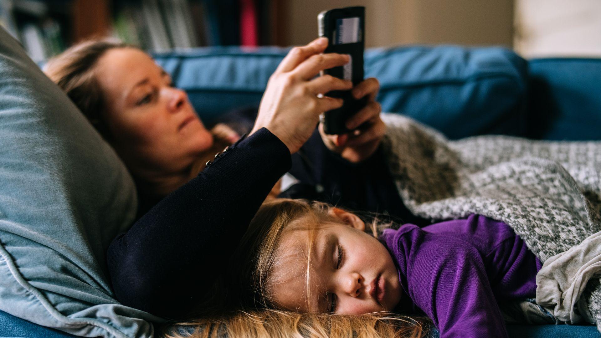 A young girl in a purple sweatshirt lies next to a woman in a black jumper on a couch. Both are covered in blankets 