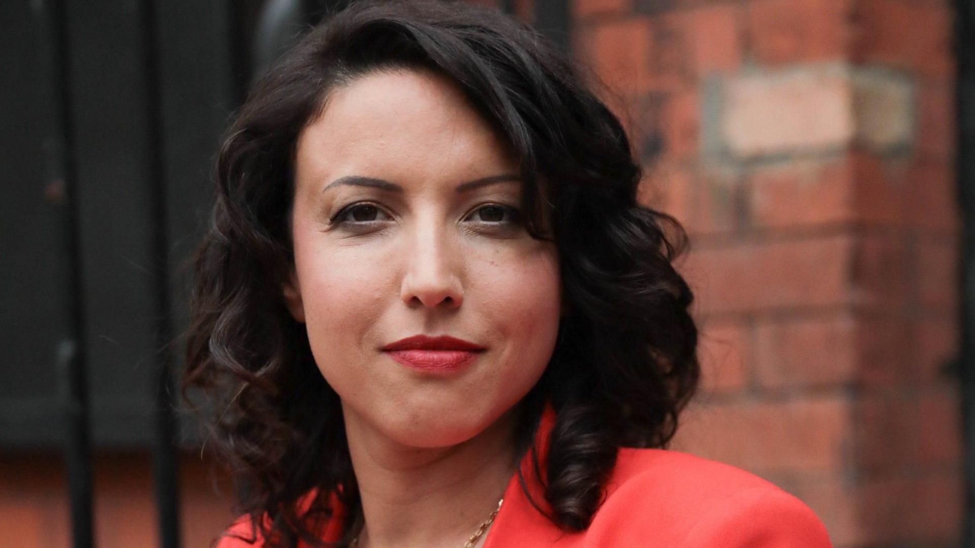 Victoria looking straight at the camera in business attire of a bright peach blazer, with dark shoulder length hair, wearing red lipstick and a gold chain.