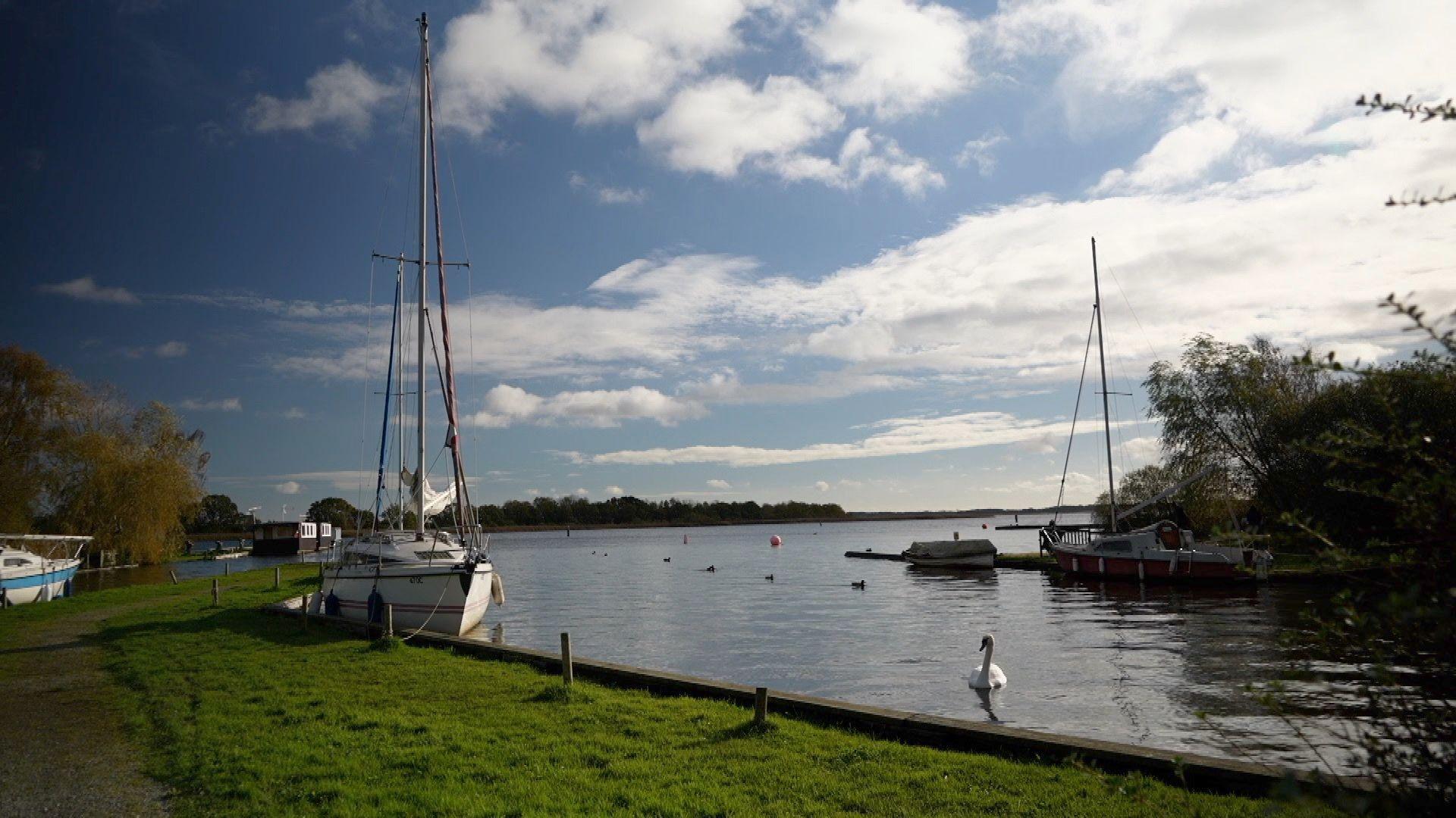 Four small boats are shown moored at the side of a waterway leading into a large broad, with grass banks lined by trees, swans and ducks in the water and a wooded area on the horizon.