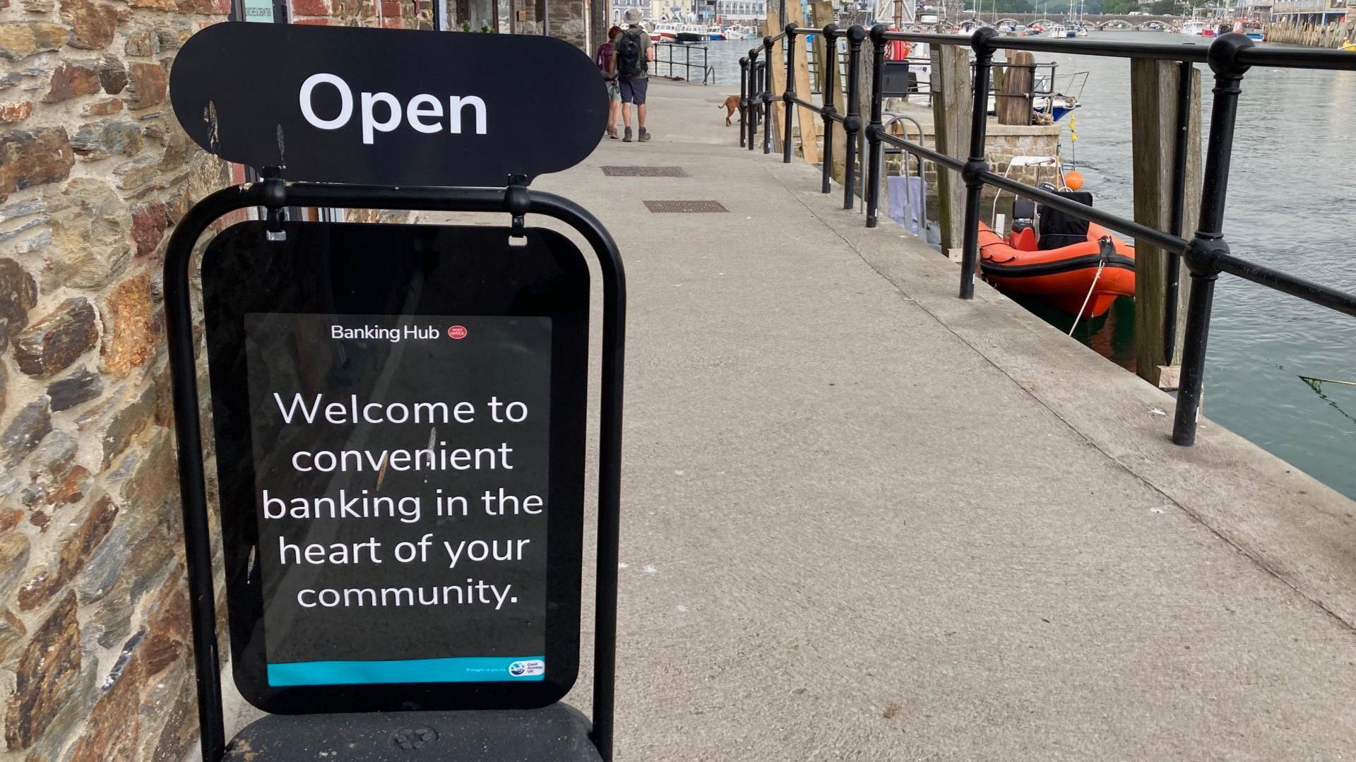 A sign that reads "welcome to convenient banking in the heart of your community" on a quayside pavement outside a banking hub.