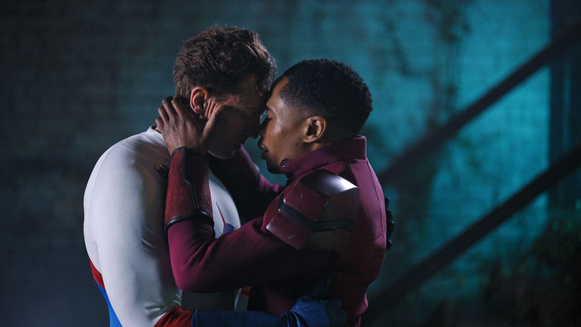 Two men in superhero costumes - one white the other wine-coloured - embrace each other affectionately, against a dark blue background.