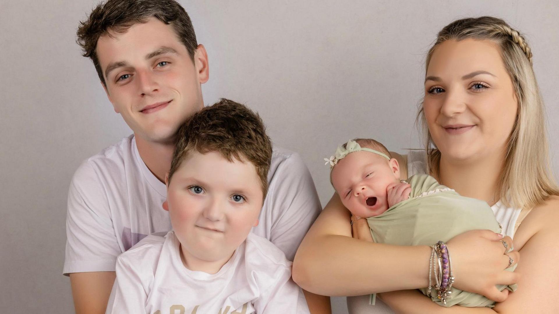 A couple, their five-year-old son and their baby daughter pose for a family portrait in front of a grey backdrop. The dad, on the left, has short brown hair and wears a white T-shirt. His son sits in front of him and has short brown hair and a matching T-shirt. The mum, on the right, has long blonde hair, partially plaited, and wears a white top and gold and purple wristbands. She is holding a baby girl who is wrapped in light green muslin and wears a green headband.