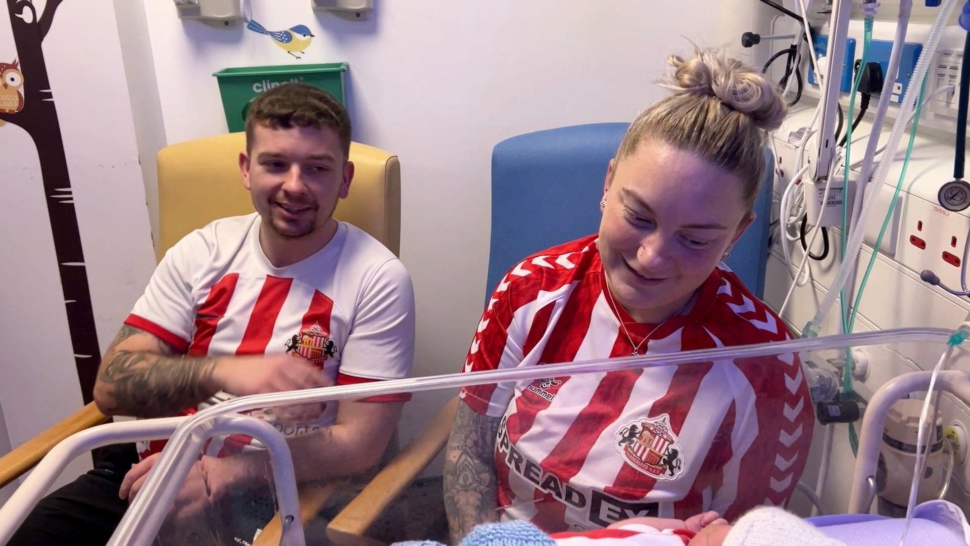 Parents Terri and Dalton Hodgen, who are both wearing the red and white Sunderland tops and are looking down and smiling at their baby. The child is wearing a wool hat and its holding its hand to its face.