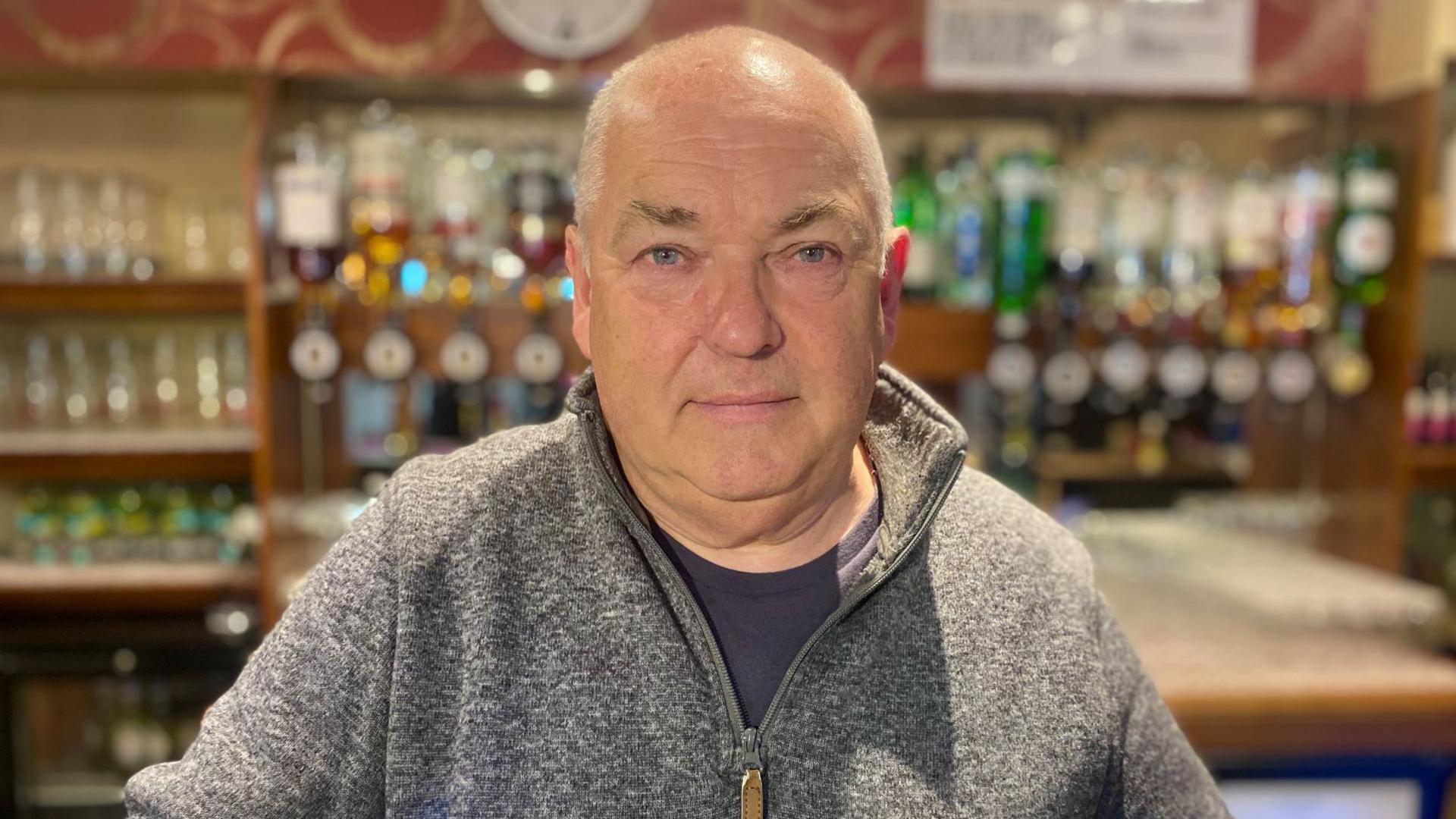 Mike has close cropped white hair. He is wearing a grey fleece over a dark grey T-shirt, and is standing in front of drinks at the bar of the Legion Club.