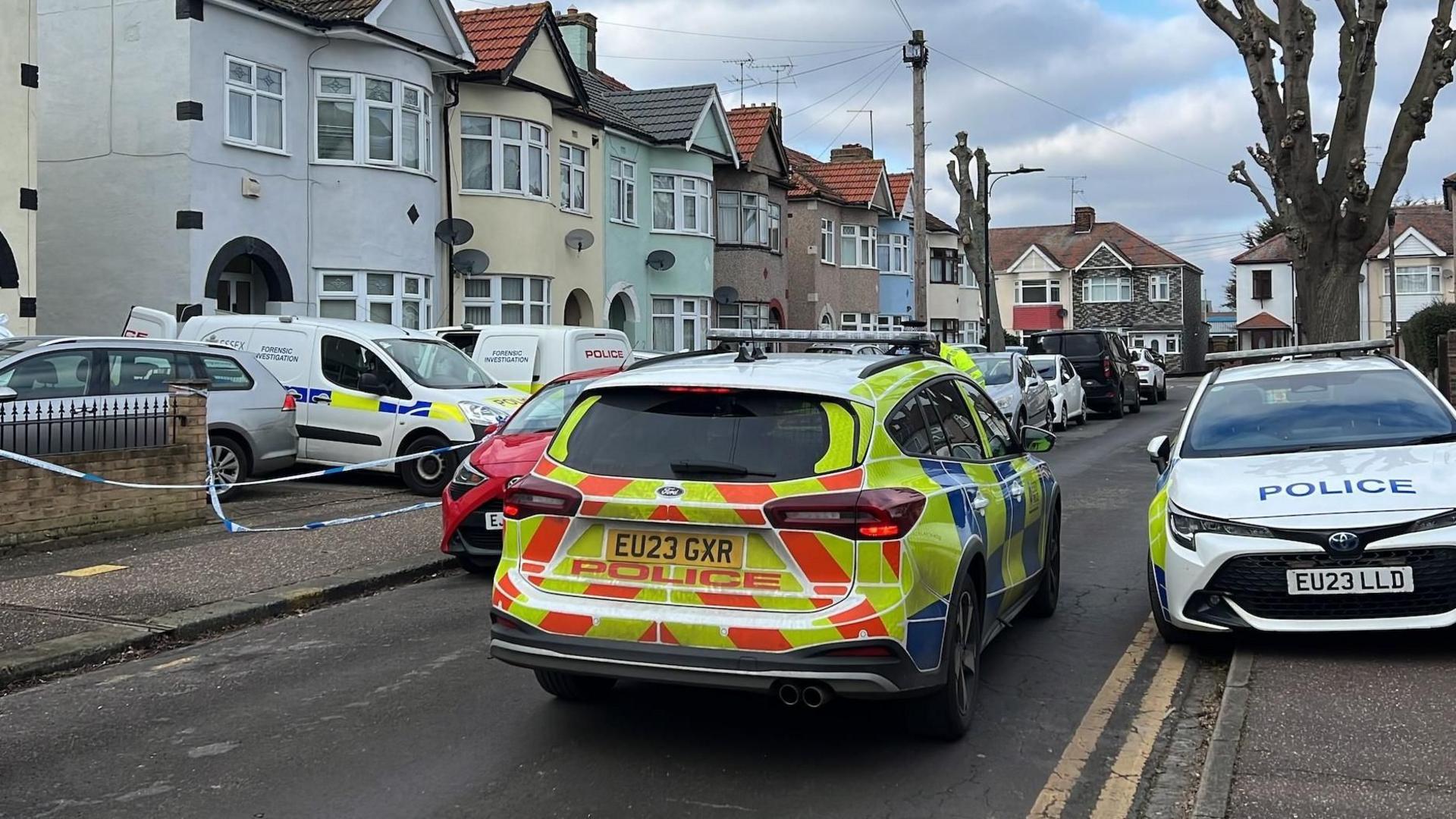 Two police cars in a residential street. There are a row of houses to the left and a tree to the right. Several police forensic vehicles are outside a house.