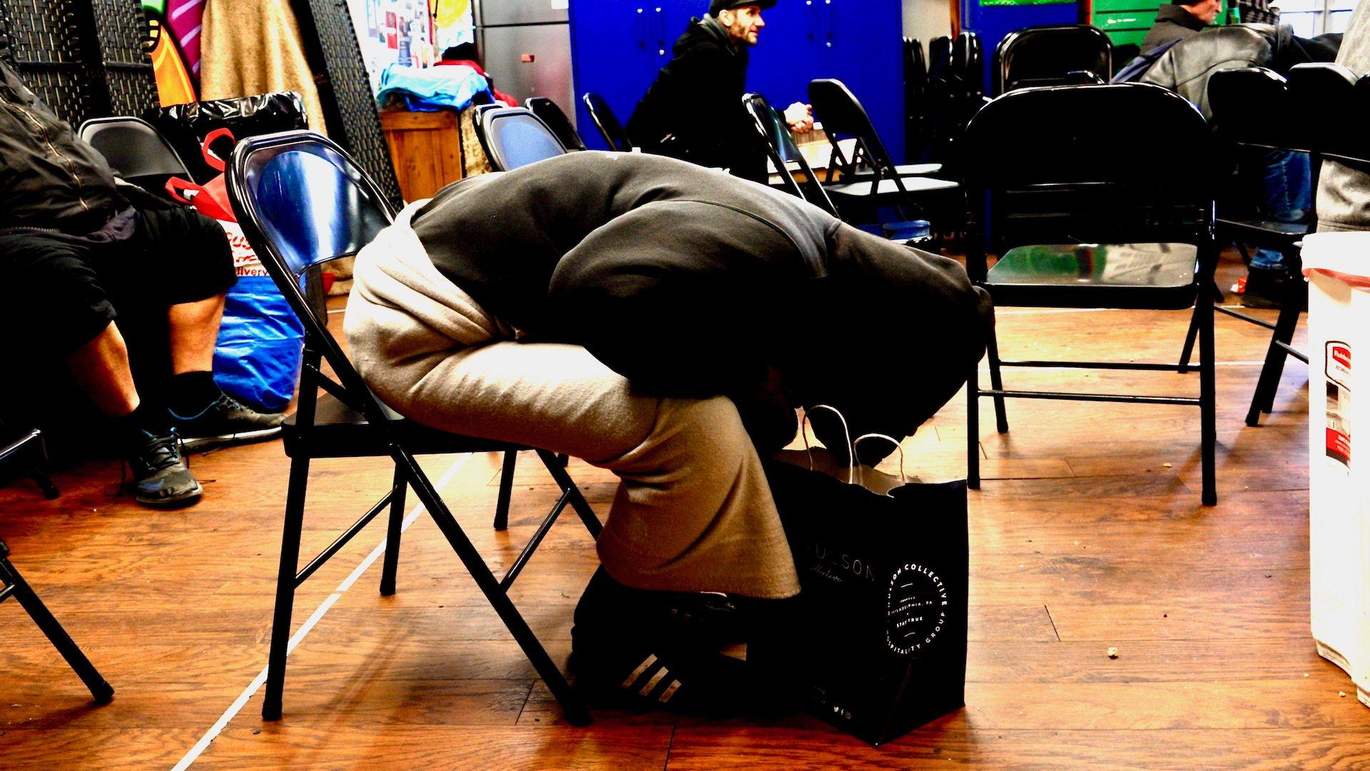 A person wearing a black hoodie and khaki trousers with black trainers is slumped forward on a fold-out chair inside the busy drop-in centre, seemingly passed out. They have a paper shopping bag at their feet.