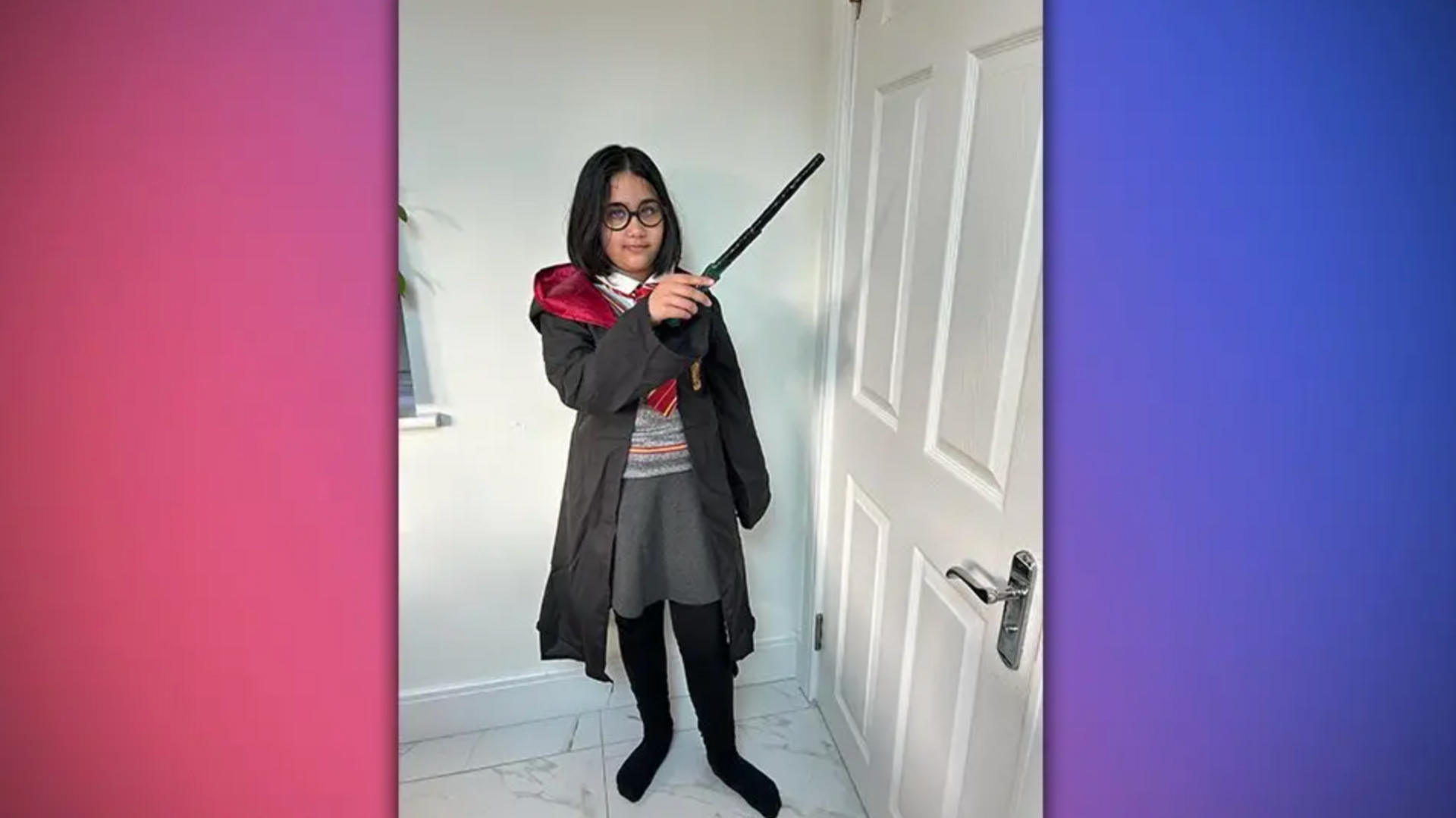 A girl with black rimmed glasses, a grey school uniform and a black a red cloak holding a wand by a white internal door