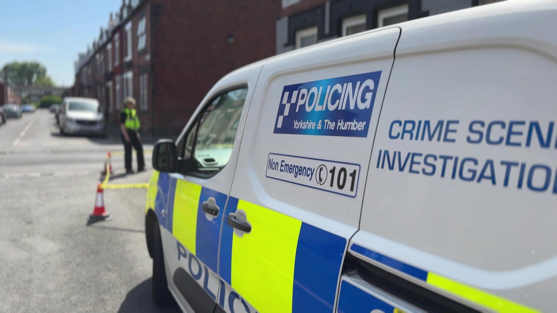 A police Crime Scene Investigation van in the foreground, parked on a residential street. In the background, a police officer can be seen stranding on the street, with cars in the distance.