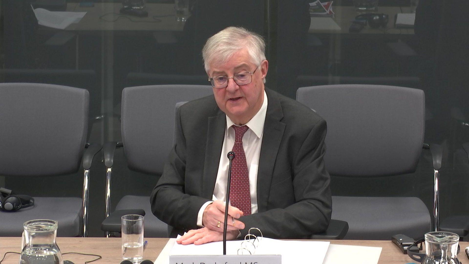 Mark Drakeford sat at a Senedd committee wearing a black suit, with a ring binder on the table in front of him and a glass of water.