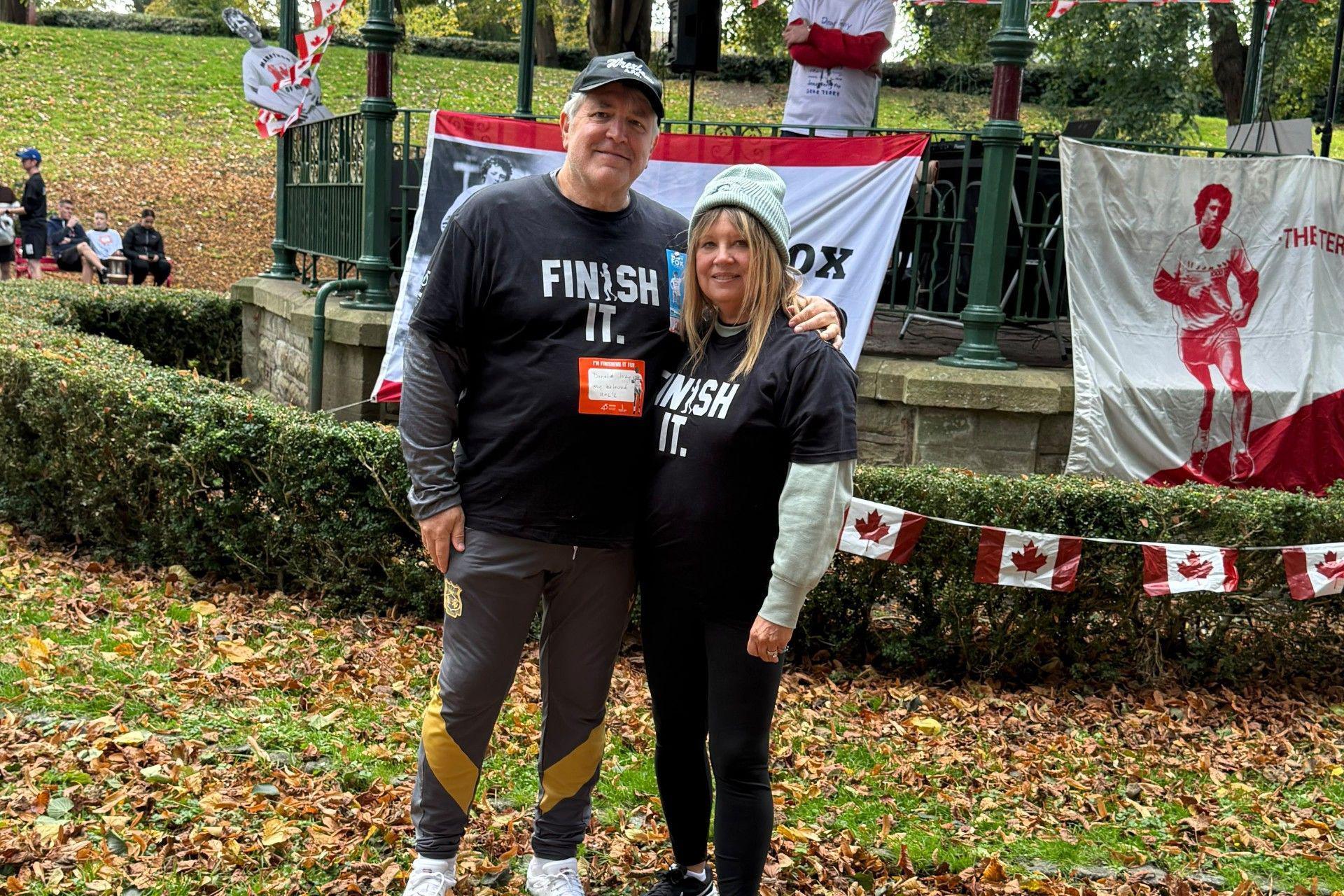 Jeff and Janice wearing black t-shirts with a logo that reads 'finish it', as well as sports leggings and trainers. They are standing in front of a bandstand in a park in what appears to be autumn with lots of brown leaves on the ground. The bandstand is adorned with bunting showing the Canadian flag and there are football related banners hanging from the railings.