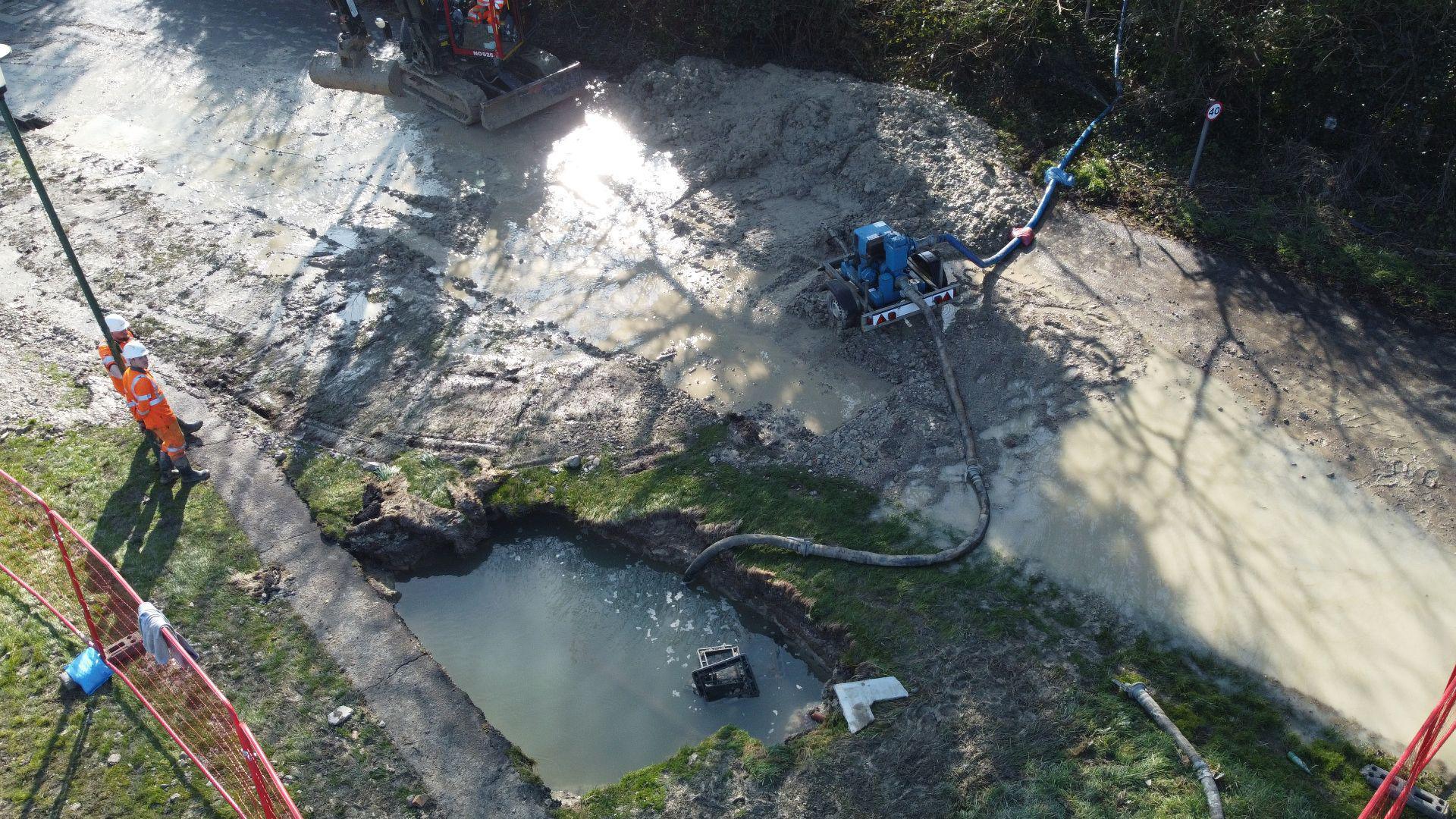 Workers stand as a large pump pumps water out of a hole in the ground. There is a red fence around the hole and some machinery nearby. 