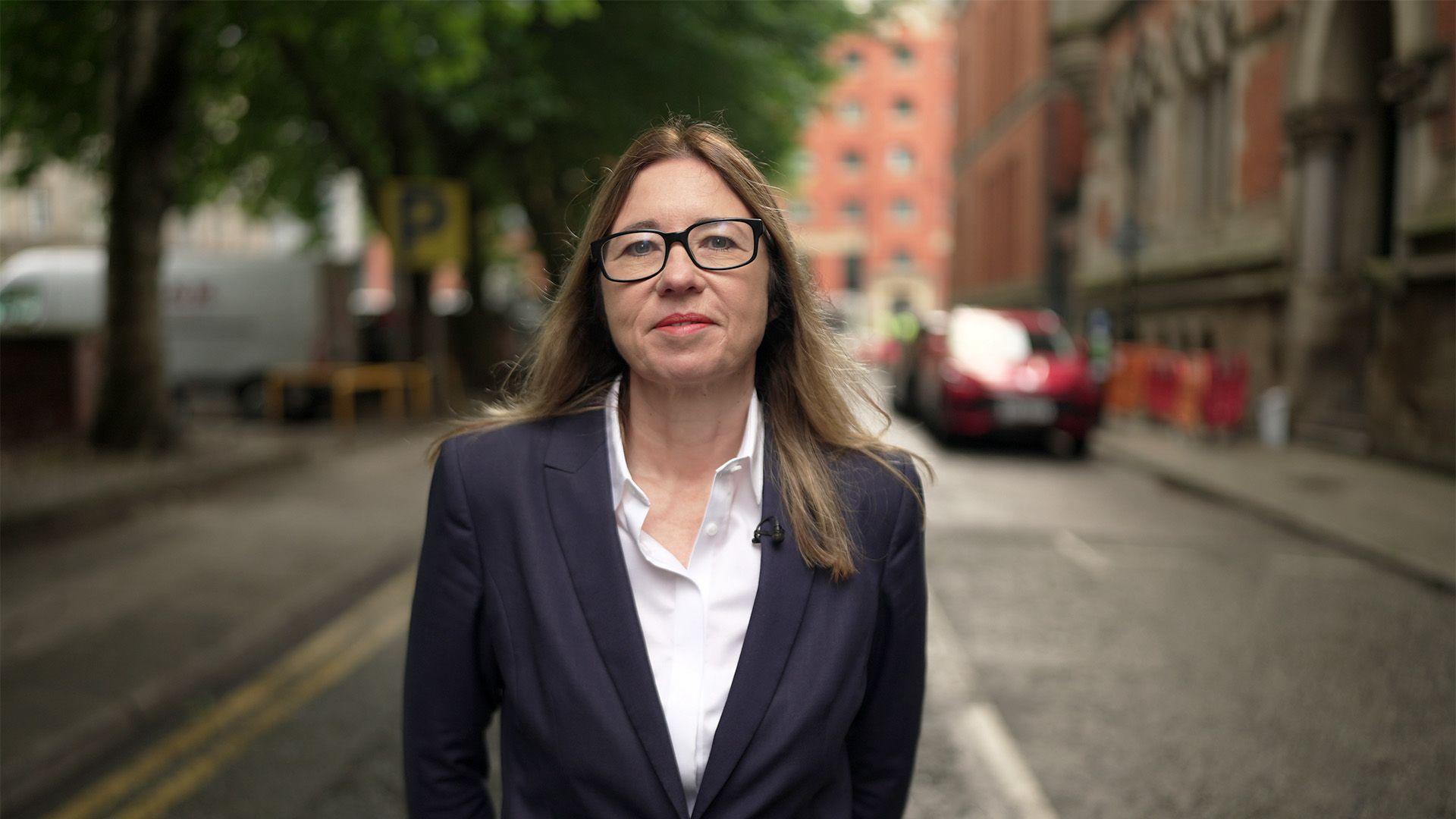 Rachel is pictured dressed smartly with long hair, wearing a dark blazer over a white shirt. She is wearing glasses and stands on a city street. The background shows trees, buildings, and parked vehicles.