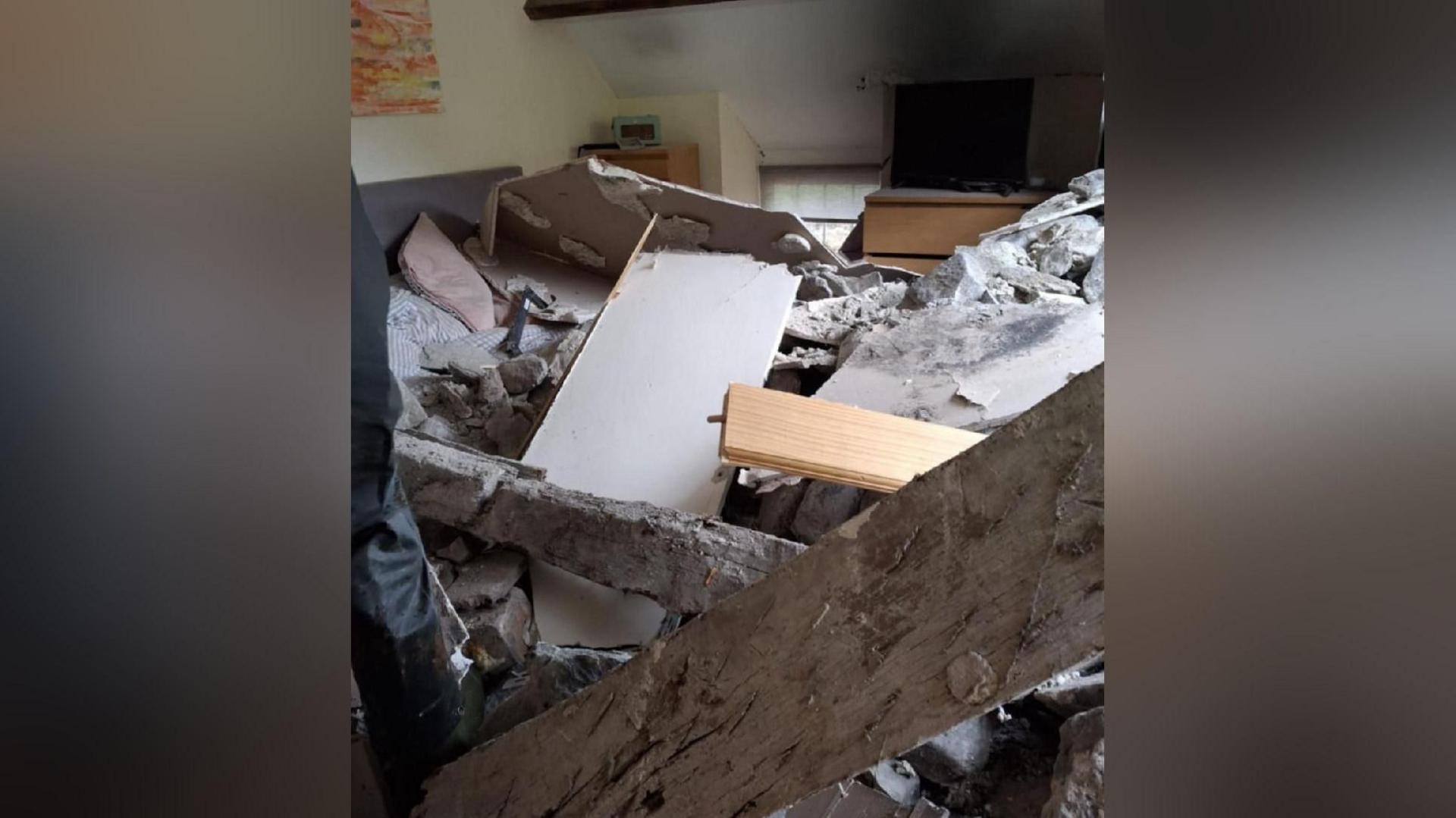 A damaged bedroom with rubble covering a bed. A large fallen wooden beam can be seen in front of the bed, while two chest of drawers and a window can be seen in the background. 