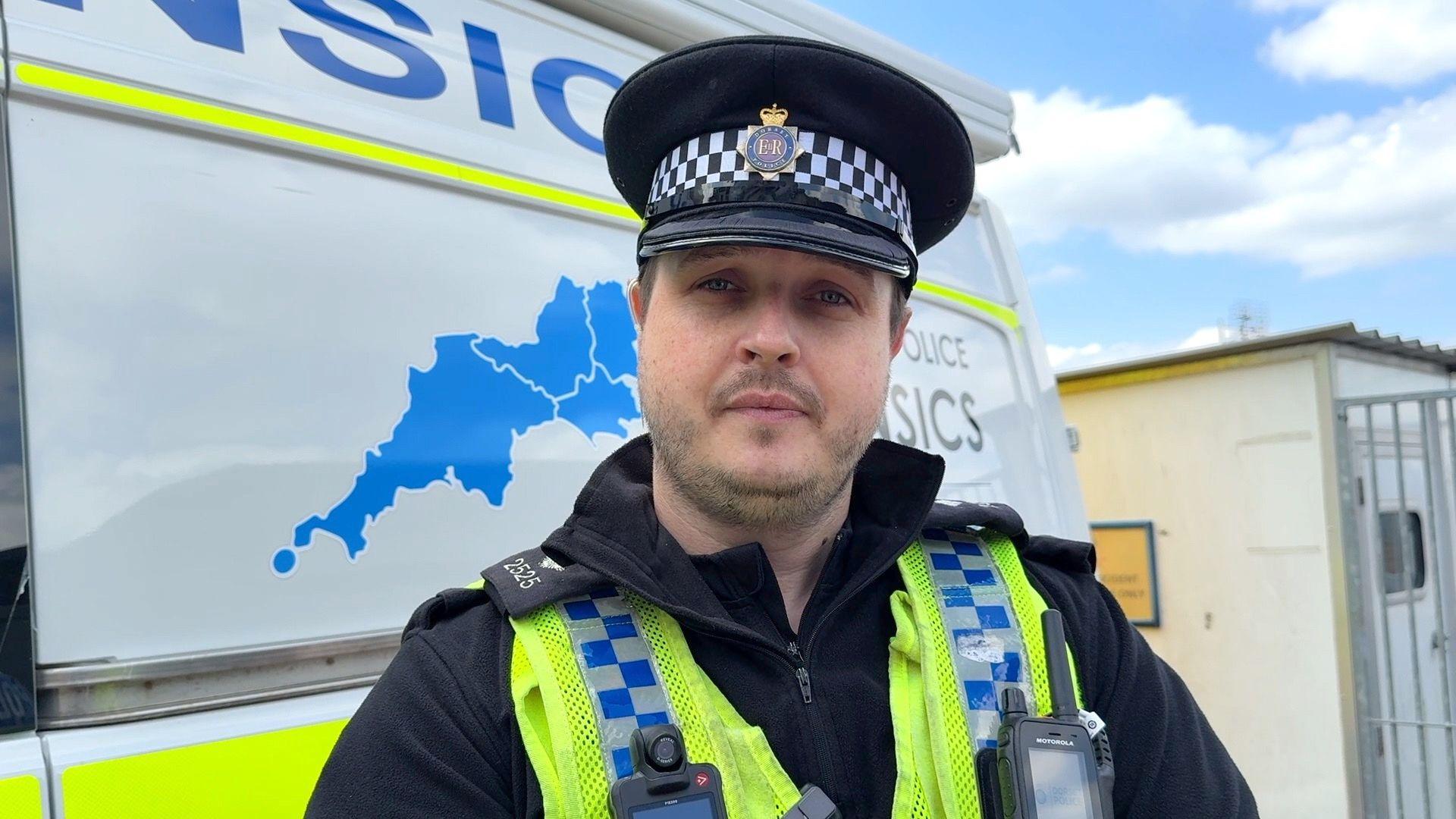 Inspector Nick Lee stood in front of a police van which has yellow stripes and a blue map of Poole on the side. He is in a black fleece which a yellow police vest on. He is also wearing a police hat. 