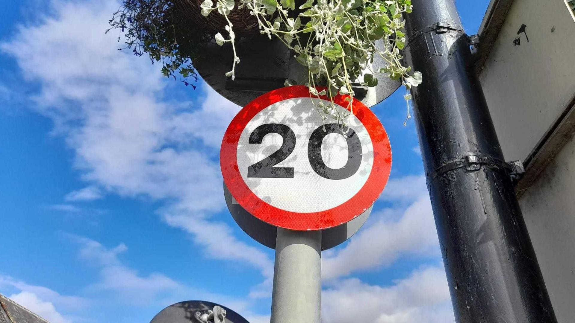 A round 20mph sign on a lamp-post. 20 is written in black letters on a white background and rimmed in red. It is underneath a hanging basket and is set against the backdrop of a blue sky with white clouds.