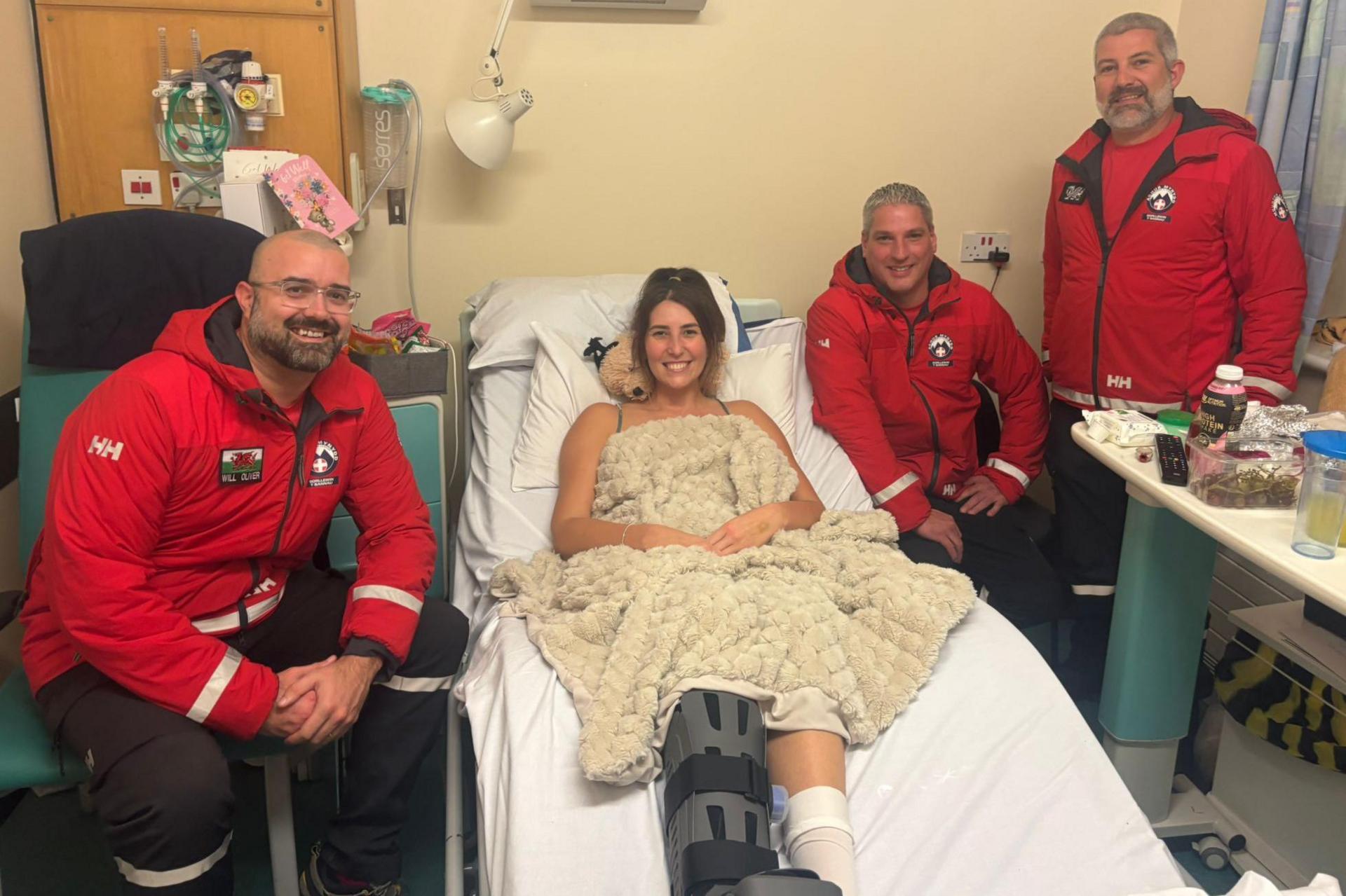 A woman with brown hair in a bun, sat in a hospital bed in a hosital room. She is covered with a blanket and one leg is in a boot, the other in a bandage. There are two men in red jackets stood beside her, with a tabke covered in drinks and snacks in front of them, and another man in a matching red jacket sat in a chair on her other side. All four people are smiling and looking at the camera.