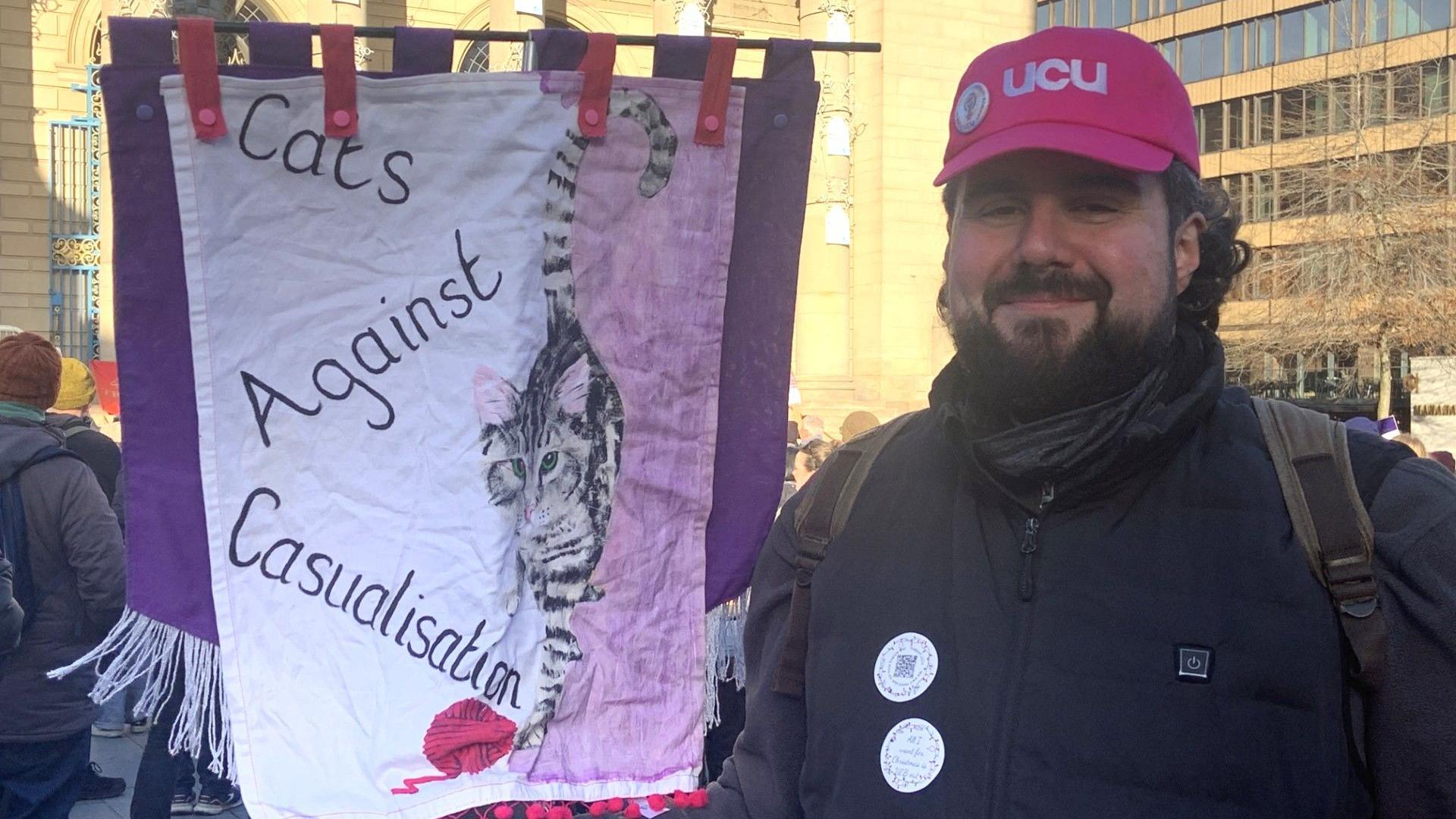 A man with a pink baseball cap and a black coat has a black beard. He is holding a pink and purple fabric banner with a cat on it which says Cats Against Casualisation