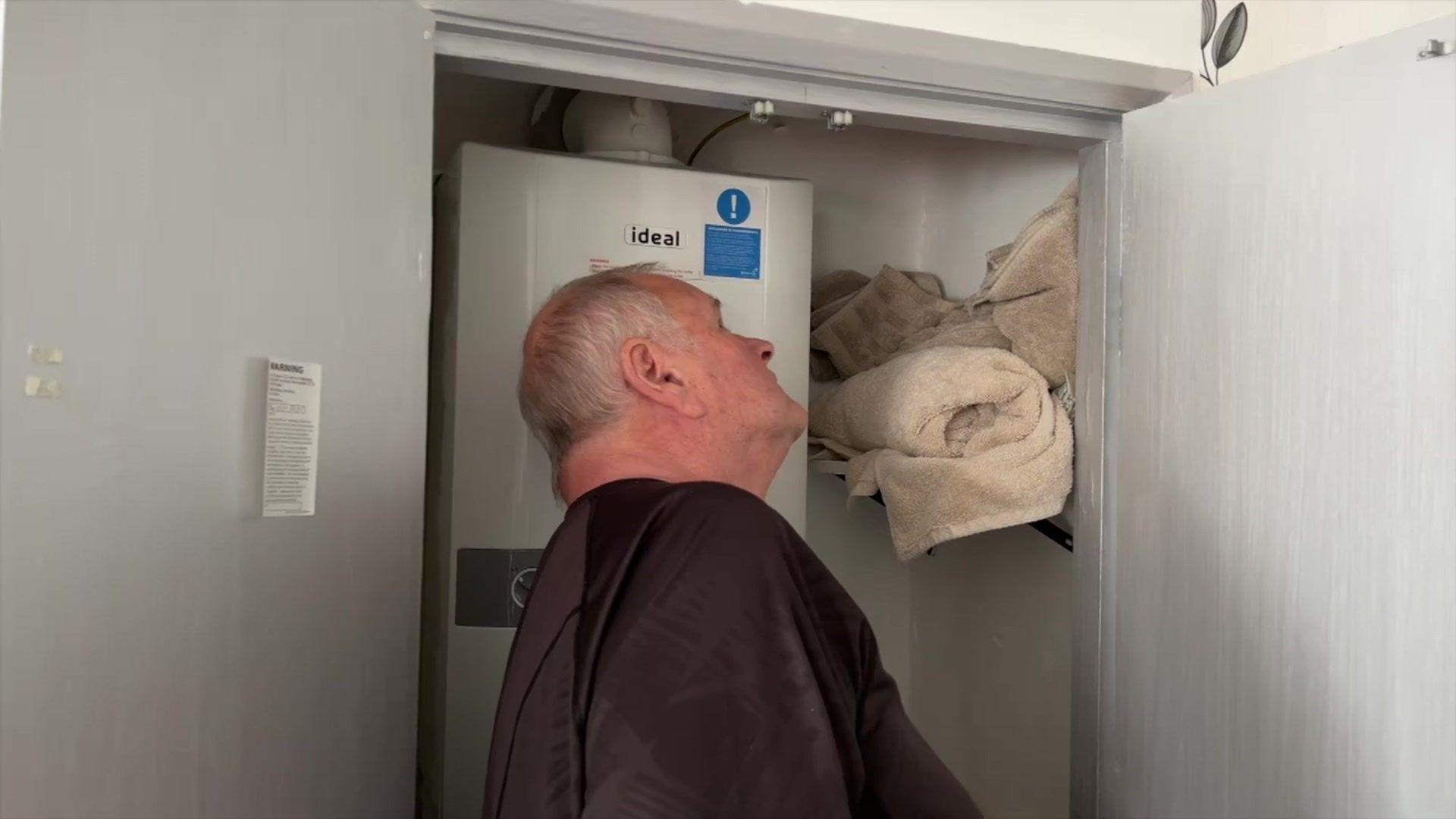 Elvet Jones looking upwards into a cupboard with a central heating boiler inside. Towels are stuffed on the top shelf to prevent water from flowing downwards