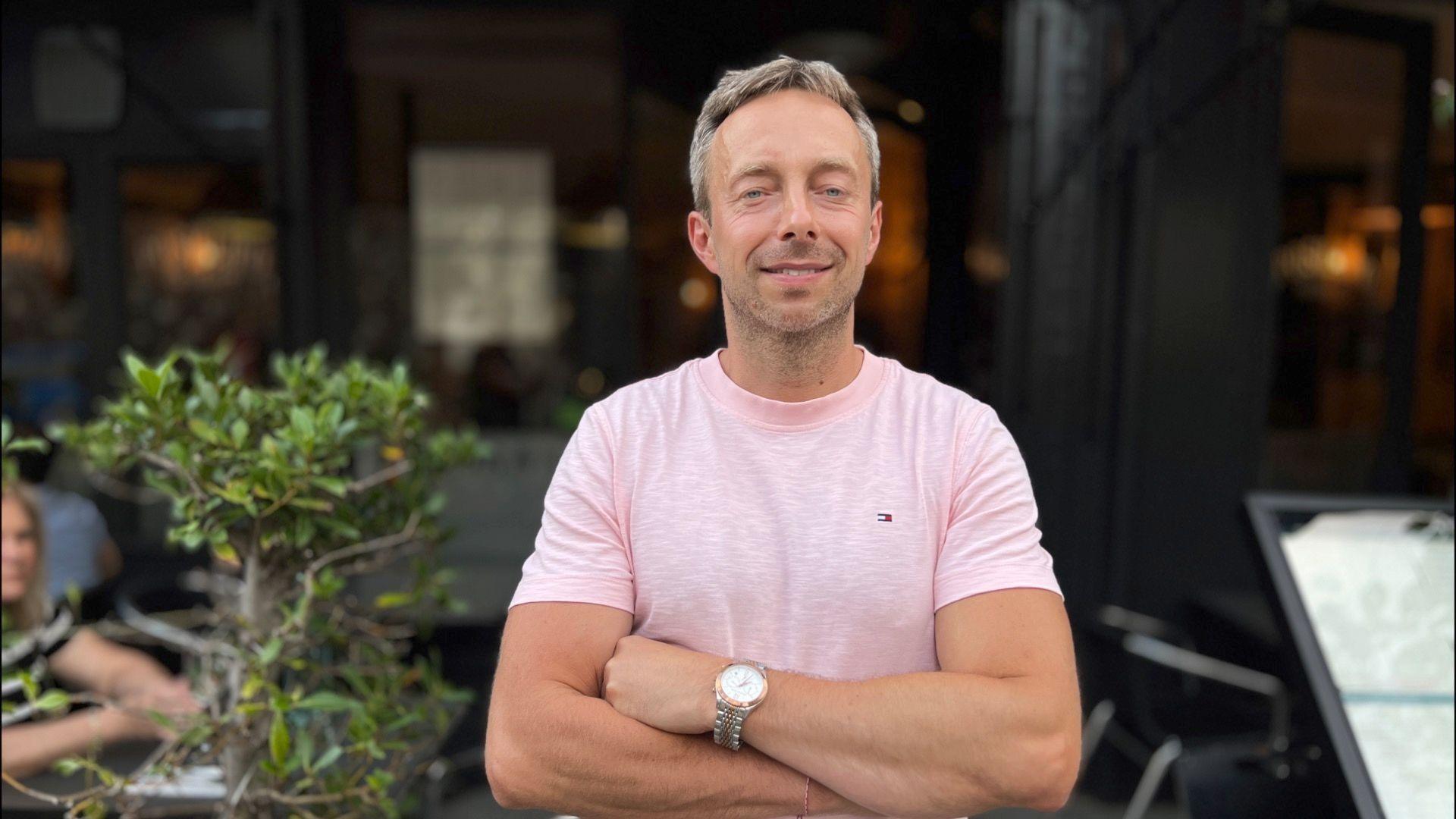 A man is standing with his arms crossed facing the camera. He has grey hair and is wearing a pink t-shirt and a silver watch. Behind him is a restaurant