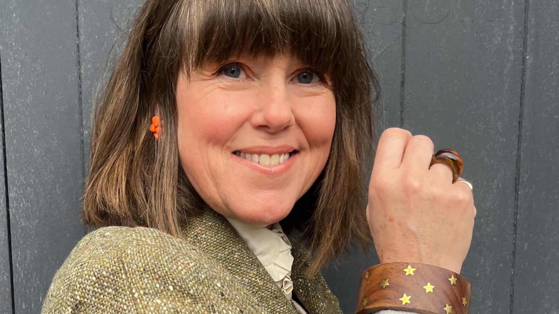 Caroline Jones is standing against a grey wooden background and is looking at the camera and smiling. She has brown hair with a fringe and blue eyes, and wears orange earrings and a chunky wooden bangle with gold stars on it. The top of a green tweed jacket and a cream blouse is just in the picture.