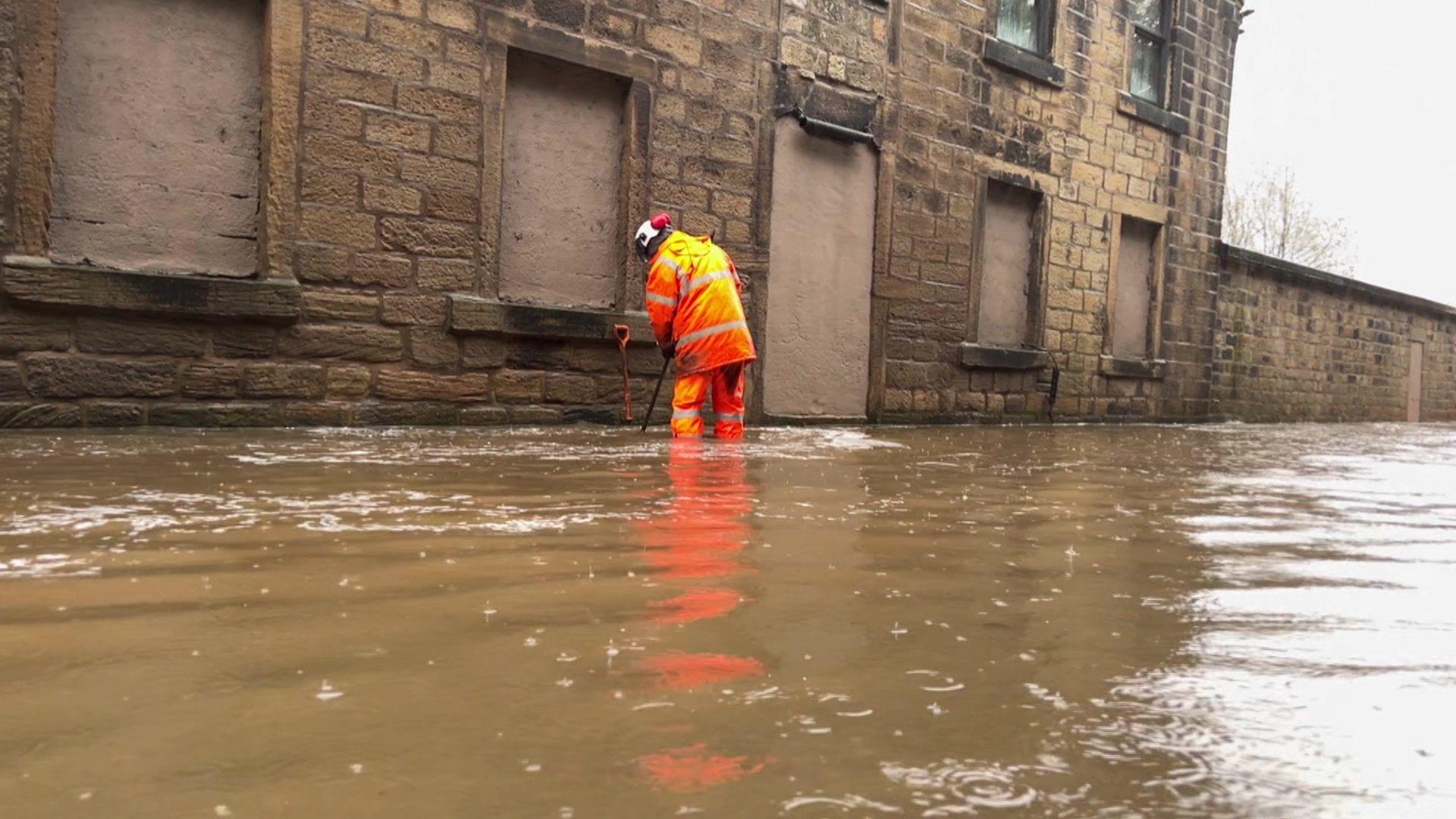 A man in orange overalls with a stick stands in floodwater next to a boarded-up property. He has his back to the camera.