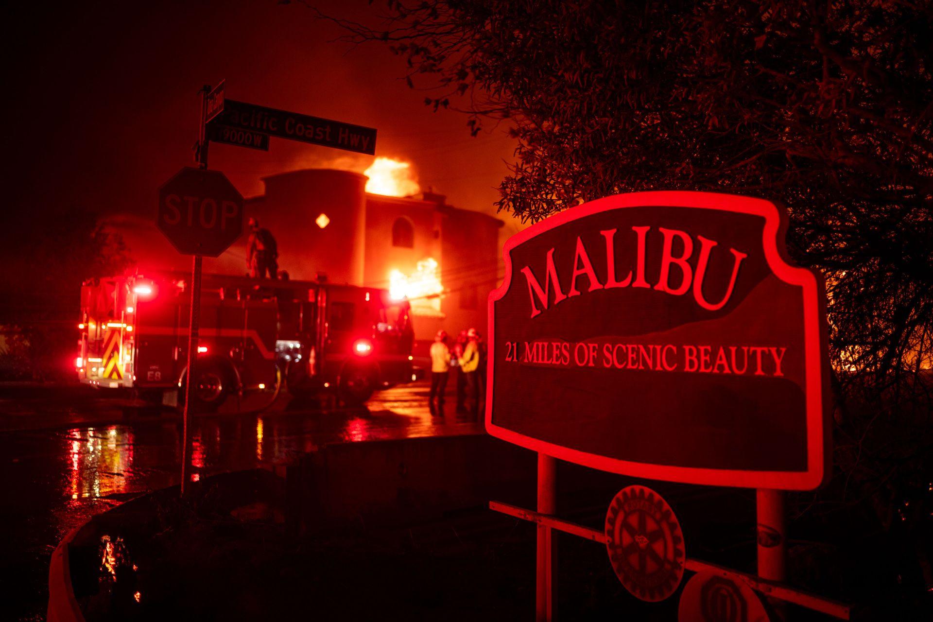 A sign saying 'Malibu: 21 miles of scenic beauty' is seen in front of a burning building with firefighters in front of it, in Malibu