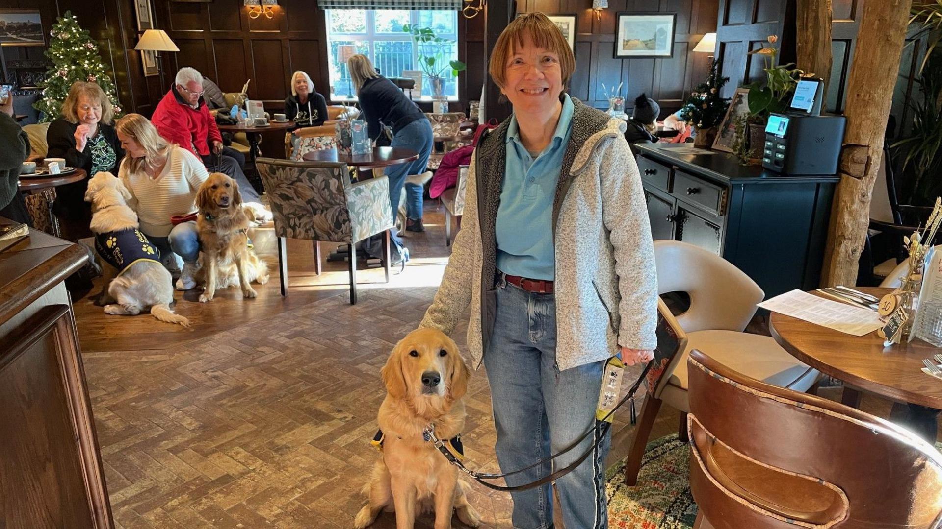 A woman in a blue polo shirt and a grey jacket stands with a golden retriever dog in a busy pub.

In the background are several other dogs and people having drinks.