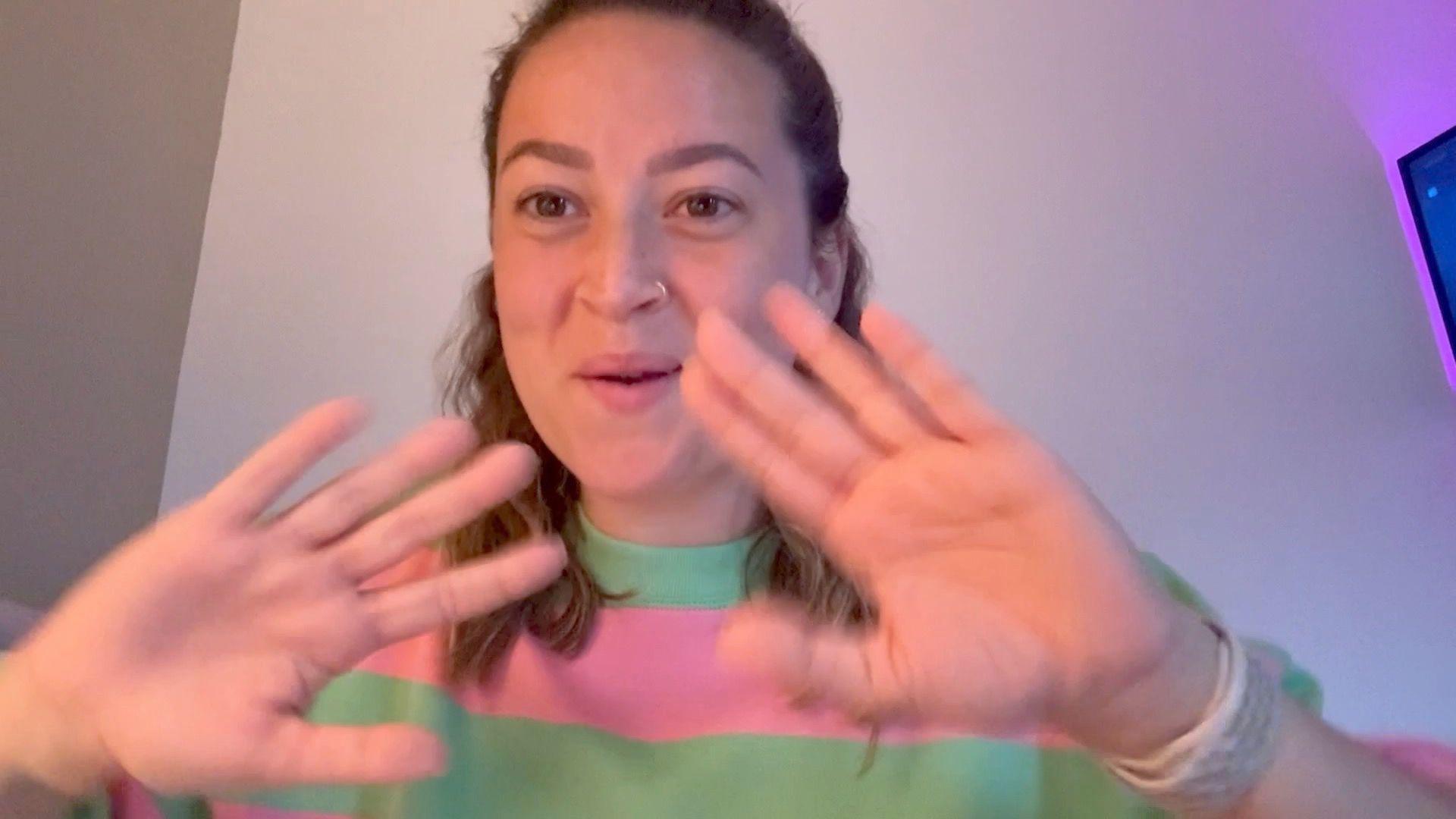 A woman with shoulder-length brown hair wearing a pink and green jumper. She has both palms of her hands facing the camera and she is deaf so is using sign language to communicate.