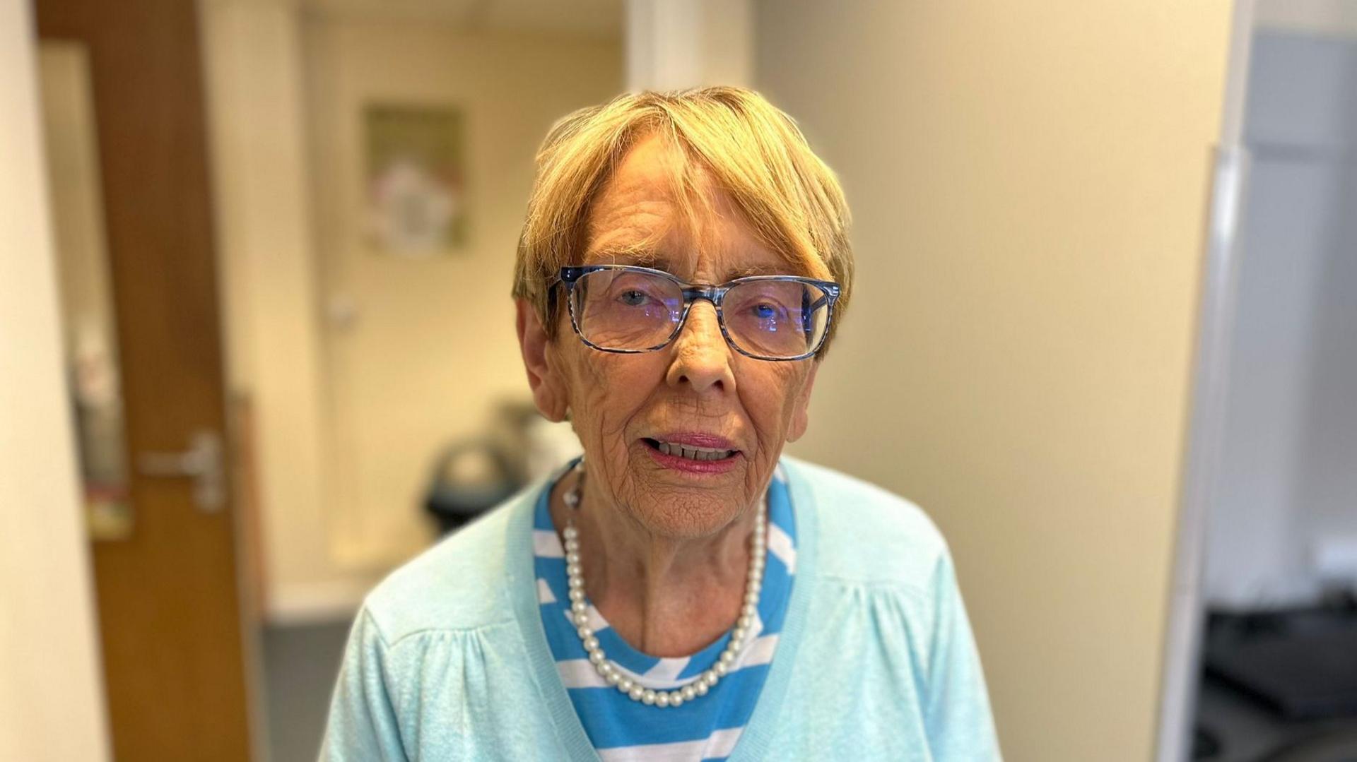 A woman with short light coloured hair is looking into the camera. She is wearing dark rimmed glasses, a blue and white striped top and a light blue cardigan with a pearl necklace.
