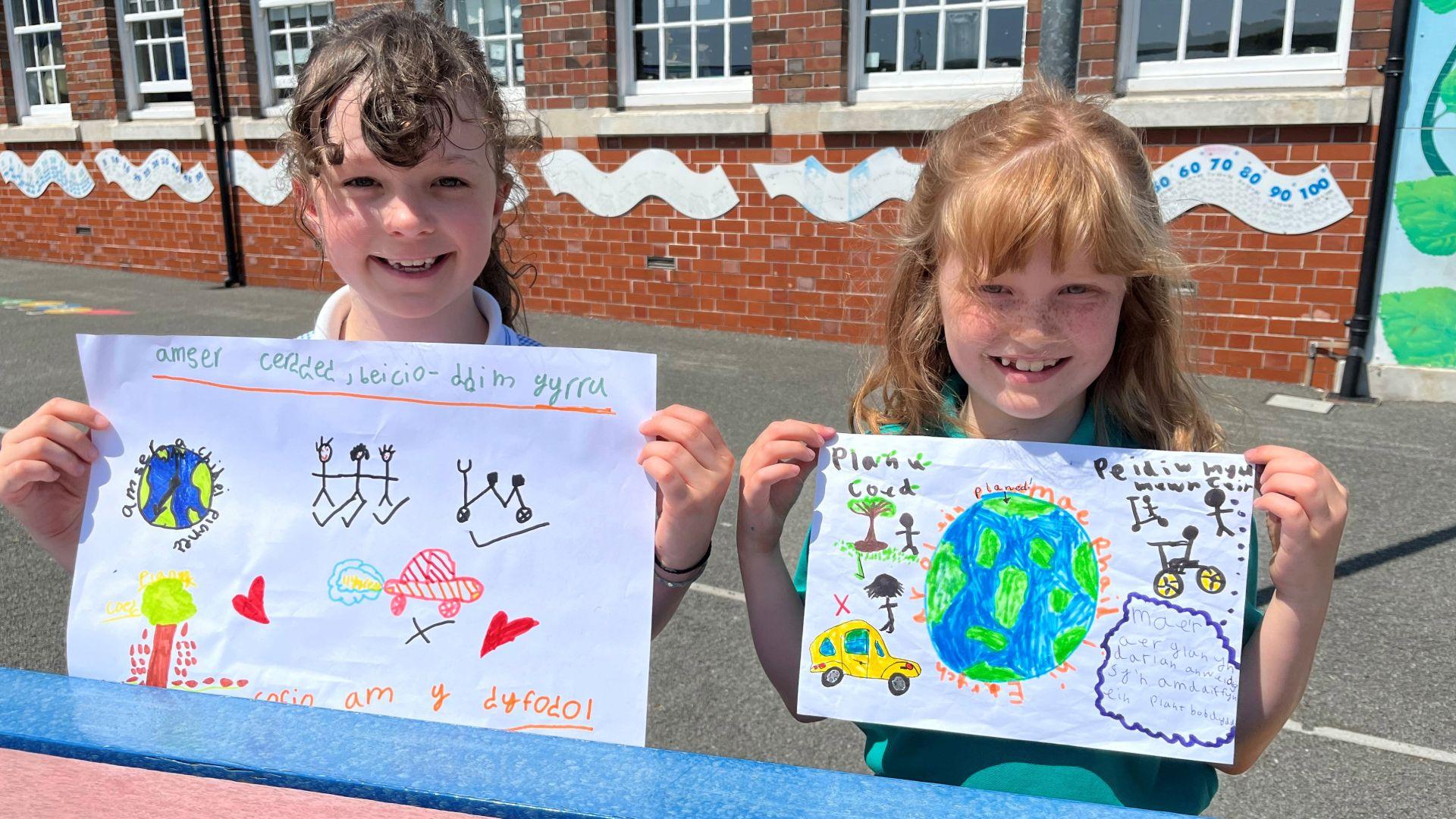 Llun o Thea (chwith) ac Amal (dde) ar iard yr ysgol yn dal eu posteri am deithio yn amgylcheddol i'r ysgol
