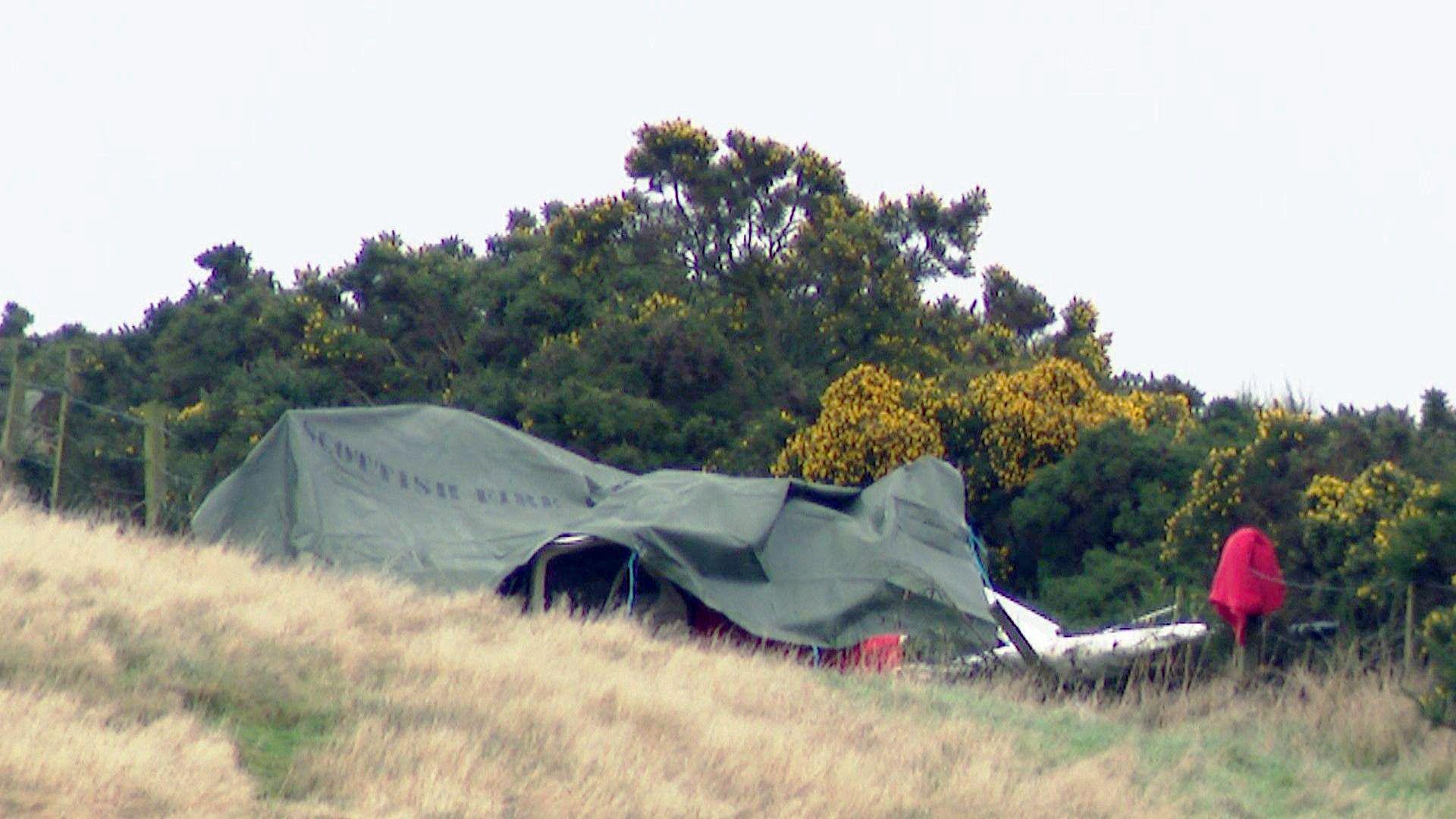 A green tarpaulin has is covering the wreckage. There are trees and bushes behind.