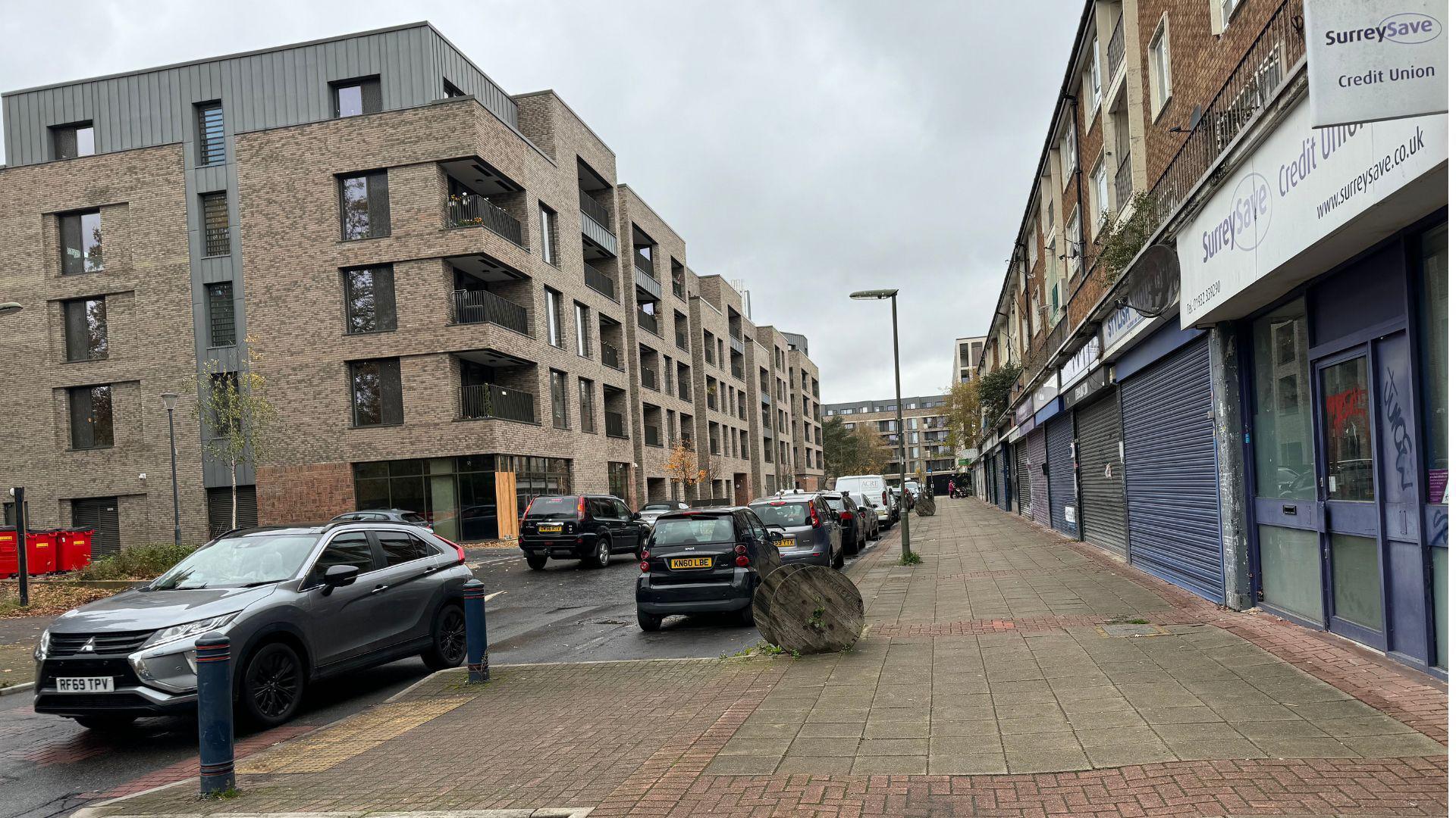 Dartmouth Avenue in Sheerwater. On one side of the road are the new flats which have been built. On the other side are older buildings with shops underneath them. One of them has a sign for SurreySave Credit Union.
