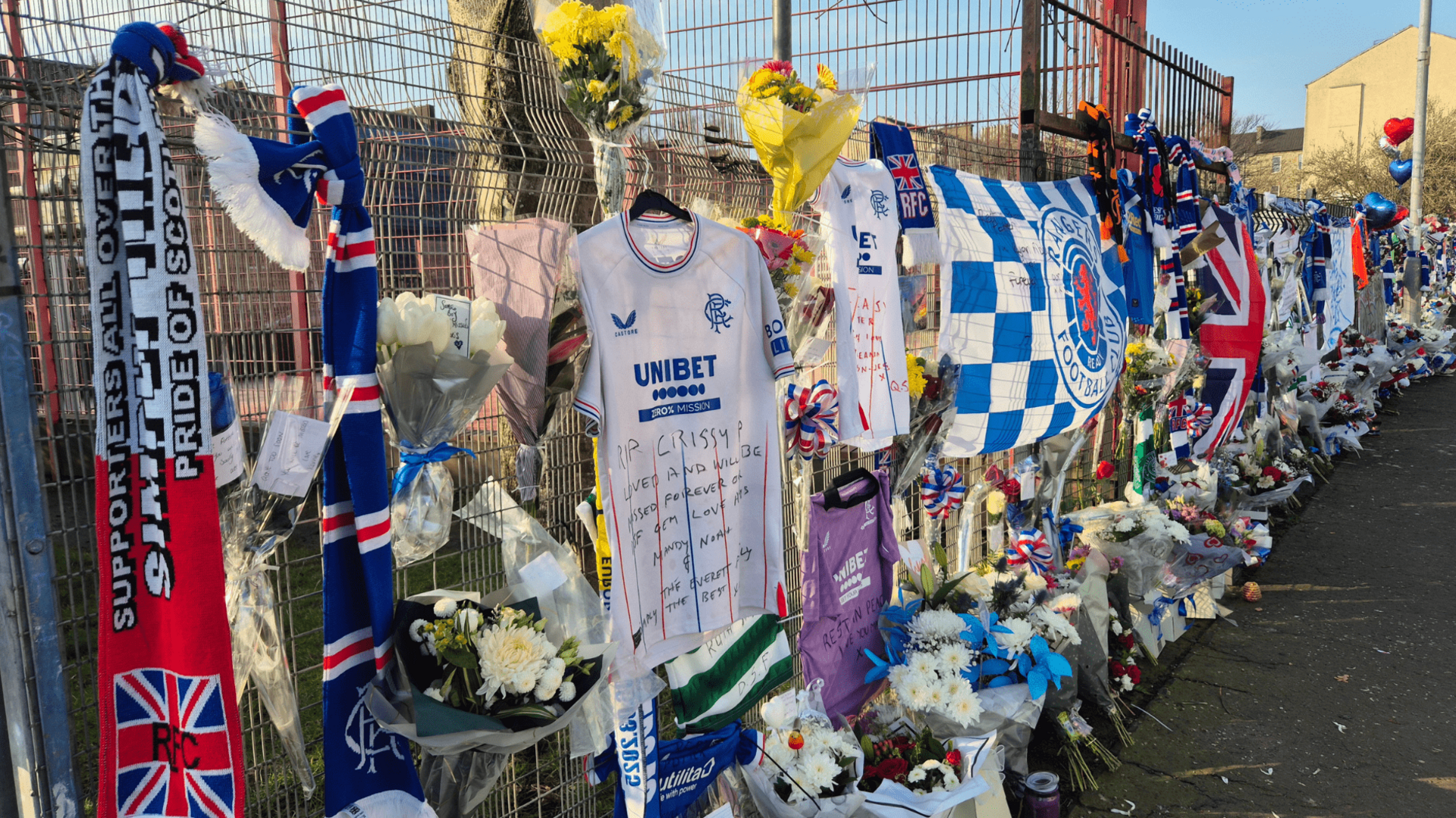 Rangers scarves, shirts and flags attached to a fence in tribute to Mr Potter. There are also several bunches of flowers and red and blue balloons.