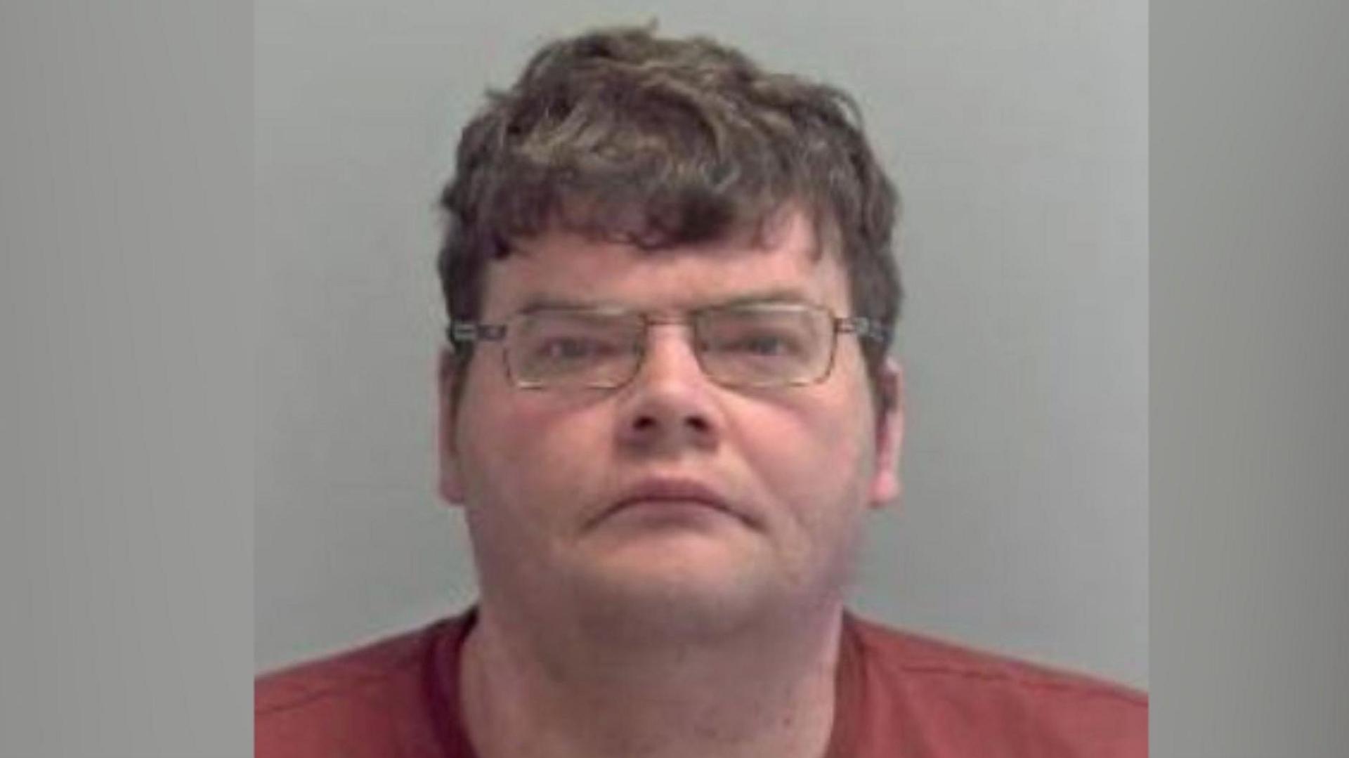 A police mugshot of Daniel Russell. He is wearing glasses and red top. 