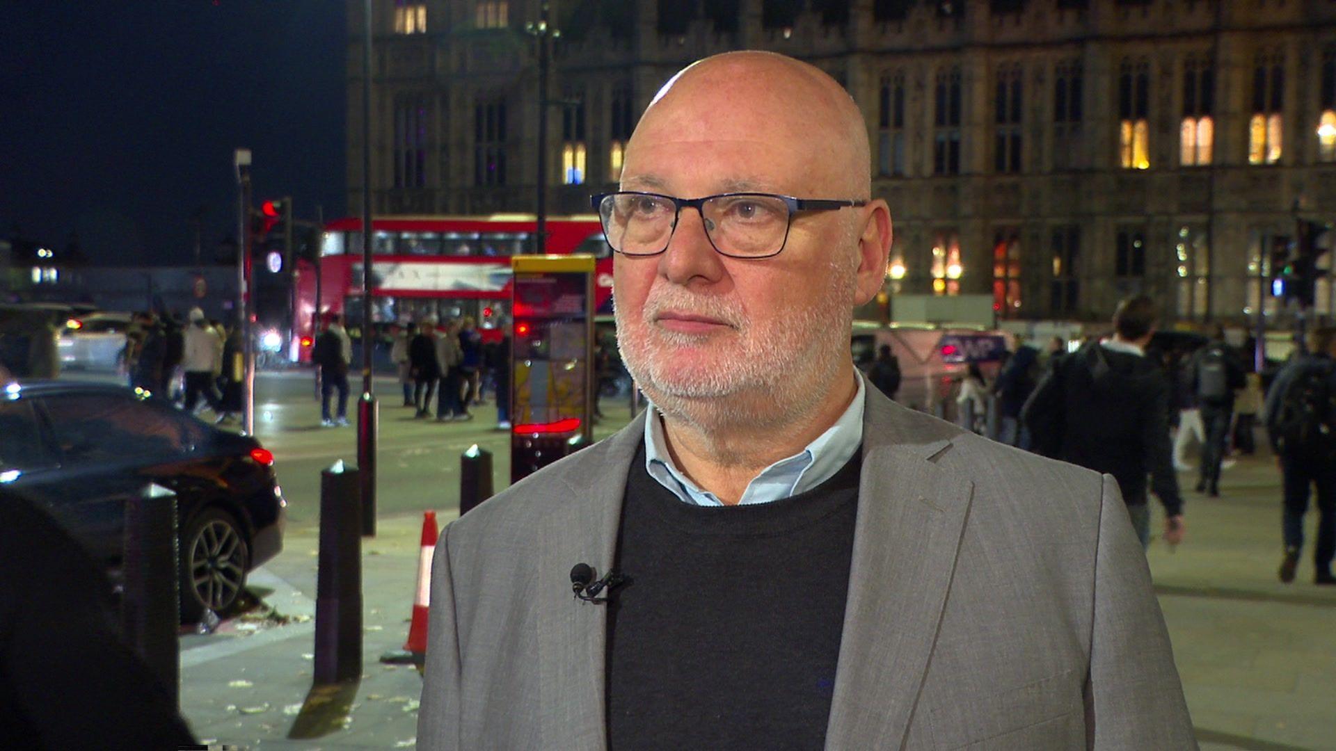 A bald man looks away from the camera. He is wearing a grey suit jacket with a navy jumper underneath and a light blue shirt. He is also wearing glasses.